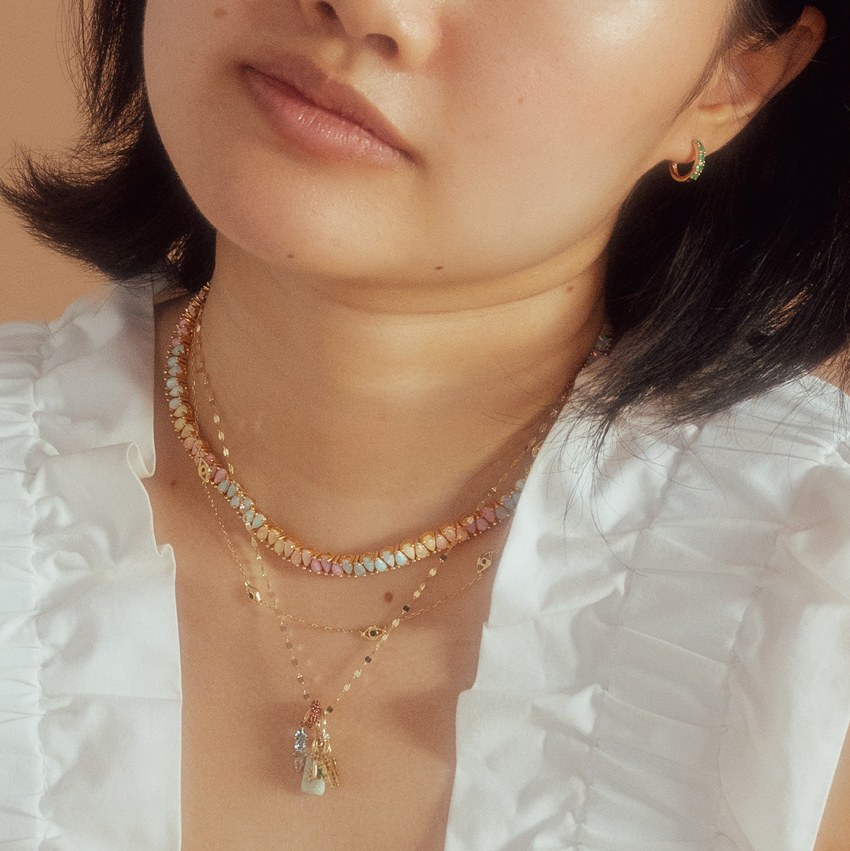 A woman wears the Opal Dancing Dreams Tennis Necklace layered with gold necklaces, a white ruffled top, and small hoop earrings, shown from the shoulders up against a neutral background.