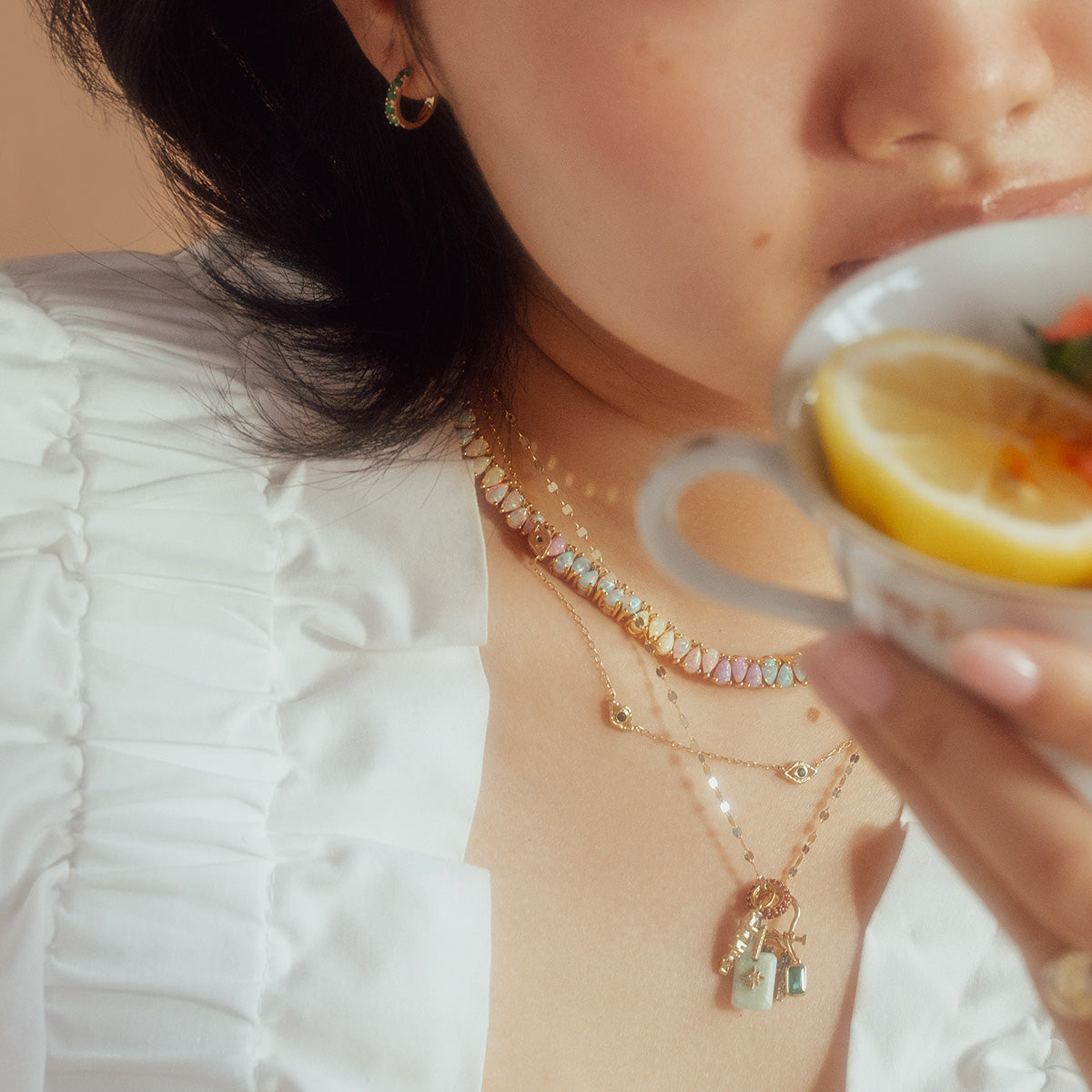 A woman in a white ruffled blouse holds a teacup with lemon slices. She layers gold necklaces, featuring the Opal Dancing Dreams Tennis Necklace, and wears small hoop earrings, her face partially out of frame.