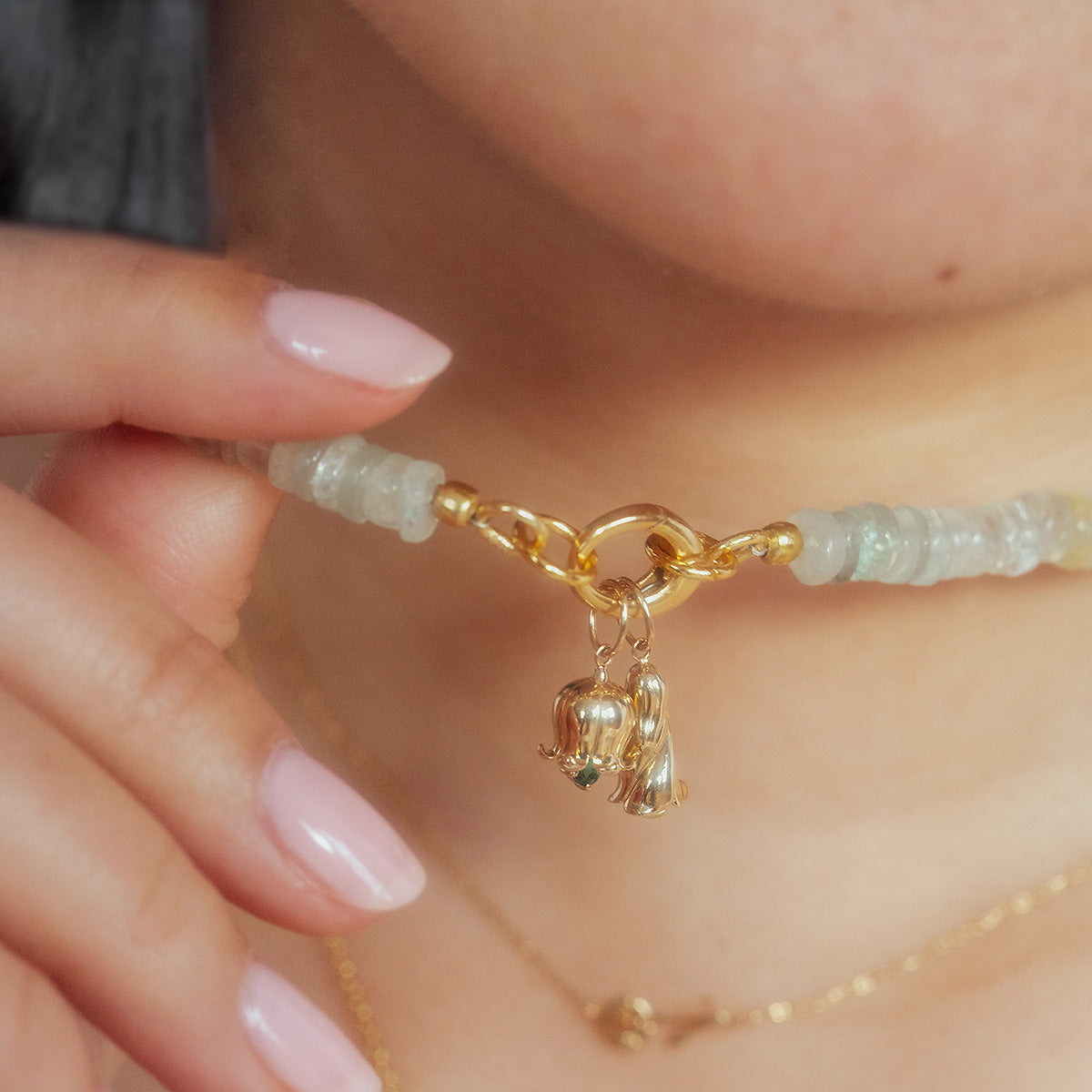 A close-up of a person with light pink nails holding a beaded necklace featuring two Solid Gold Birth Flower Bud Charms crafted in 9k yellow gold. The person also wears a delicate gold chain necklace.