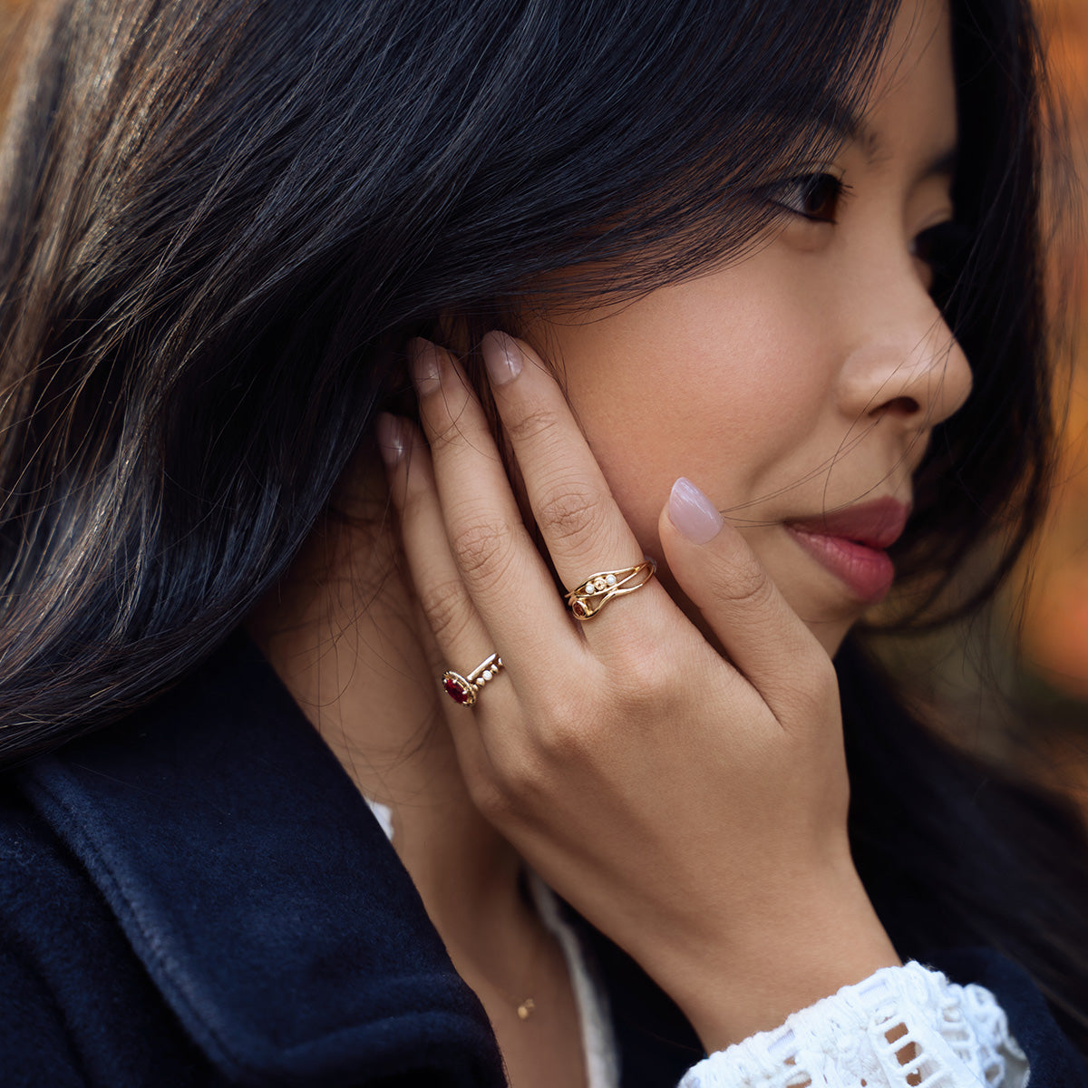 A woman with long dark hair touches her face, showcasing the 10k Lucky In Red Ruby Halo Ring on her finger. She wears a dark coat and white top, with a neutral expression and natural makeup.