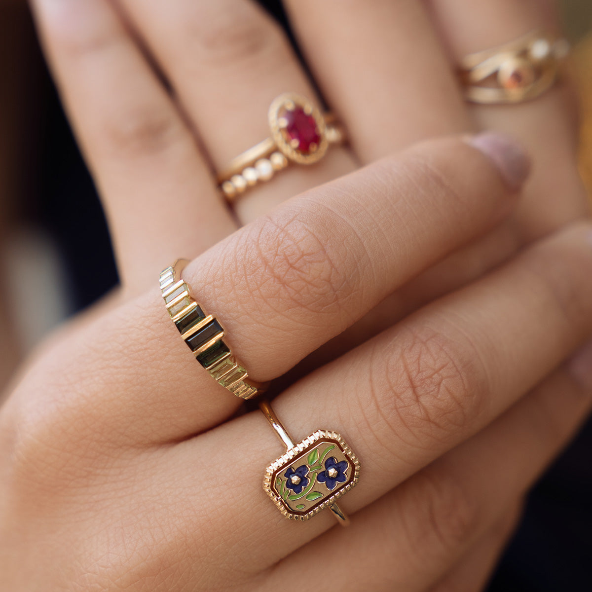 Close-up of a hand wearing four gold rings, including the 10k Green Tourmaline Skyline Band, a ring with a colorful floral enamel design, one set with a red gemstone, and two simple, elegant bands.