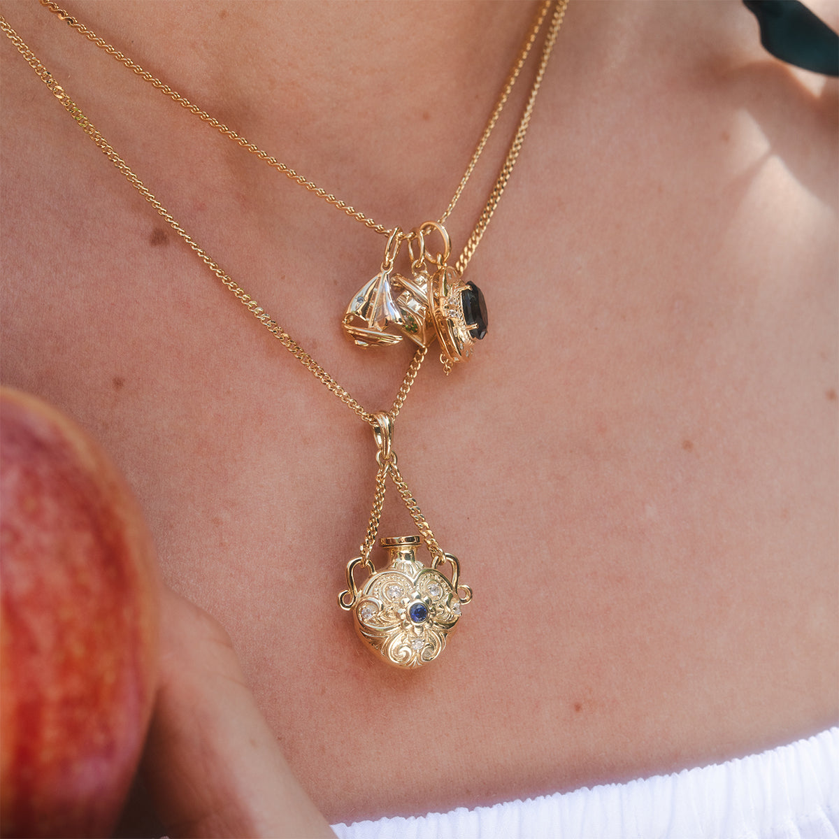 A close-up of a person wearing layered gold necklaces, including the Juliet Vial Pendant Necklace and a filigree blue stone pendant. The person holds an apple and wears a white off-shoulder top.