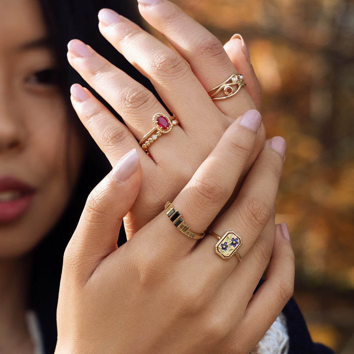 A woman with long dark hair displays her hands wearing four gold rings, including the 10k Green Tourmaline Skyline Band, each with unique gemstones and designs, set against a soft autumn outdoor backdrop.