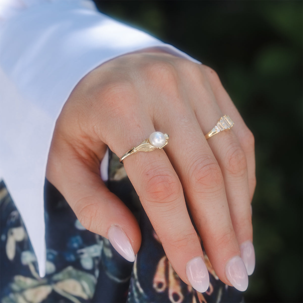 A hand with light pink manicured nails wears the Pearl Siren Statement Ring, featuring a white pearl, alongside another gold ring. The hand rests on patterned fabric and is paired with a white long-sleeved shirt.