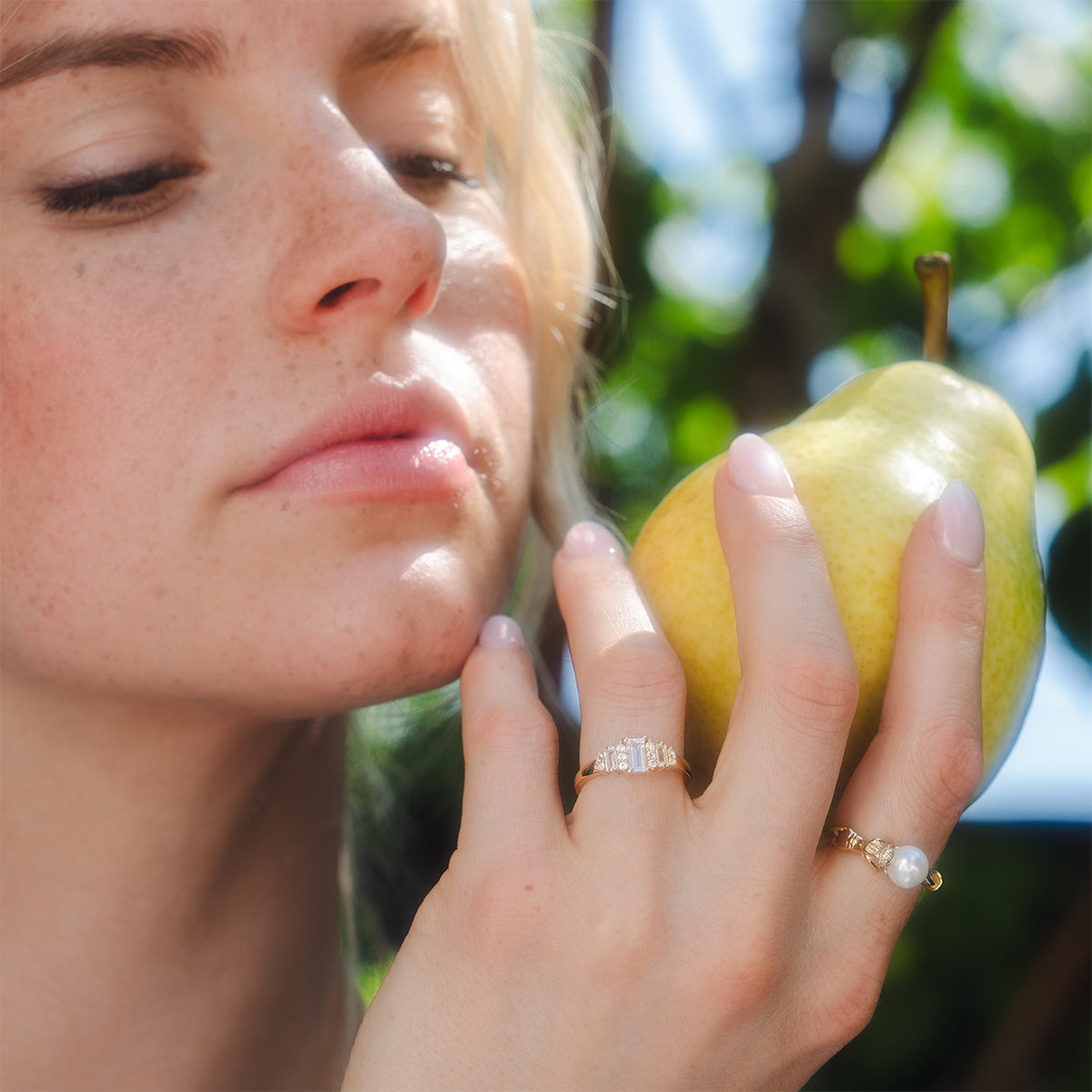 A woman with fair skin and light hair holds a yellow pear near her face, highlighting the Third Act Art Deco Ring on her finger. The rings 14k gold vermeil vintage style shines, set against a blurred background of green foliage and sunlight.