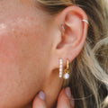 Close-up of a person’s ear wearing the Pearl Keepsake Huggie Hoops—gold vermeil earrings with small pearls—alongside a thin silver hoop and pearl drop earrings. Manicured fingers gently touch the earlobe.