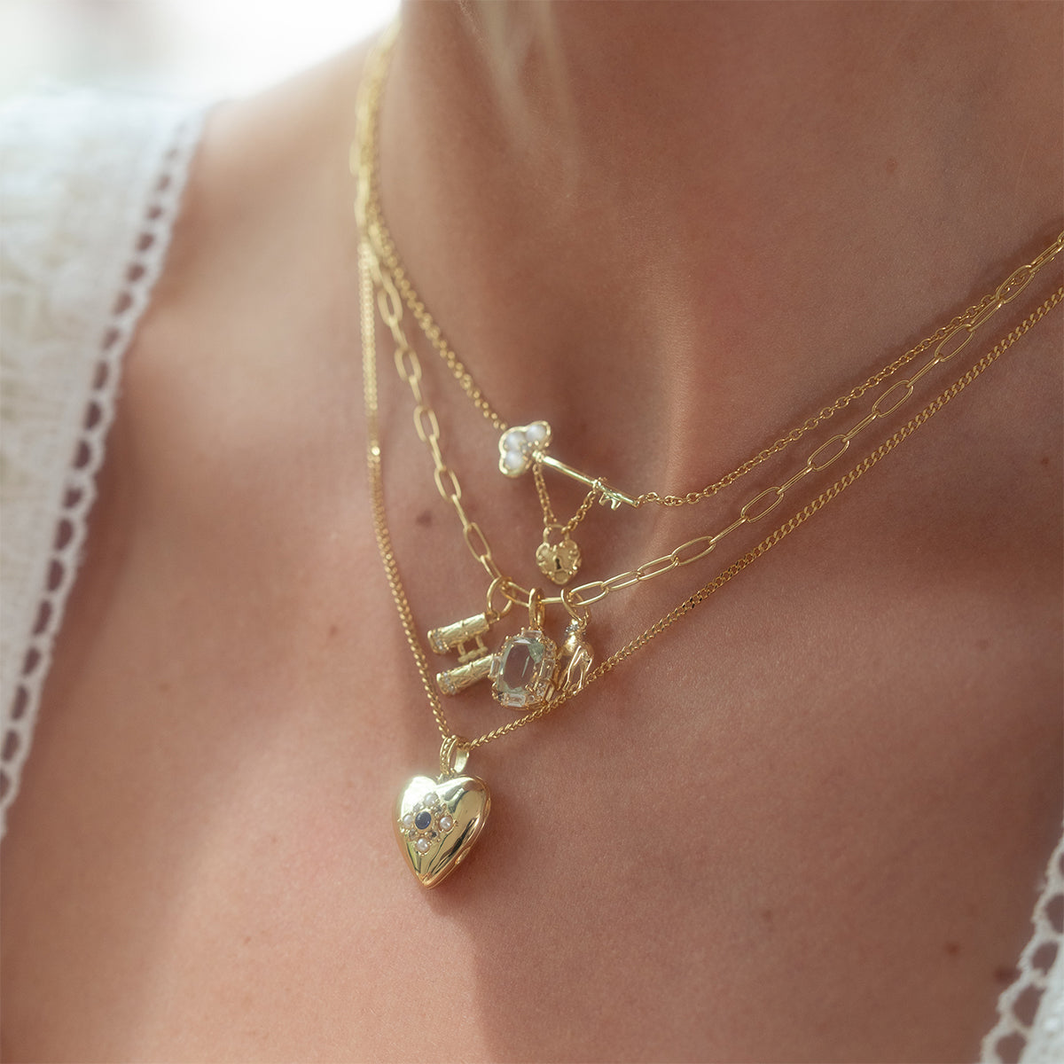 Close-up of the Vintage Inspired Trinket Charm layered gold necklaces—featuring heart locket, key, lock, gemstone, and round charms—over a lace-trimmed top for an elegant period-inspired look.