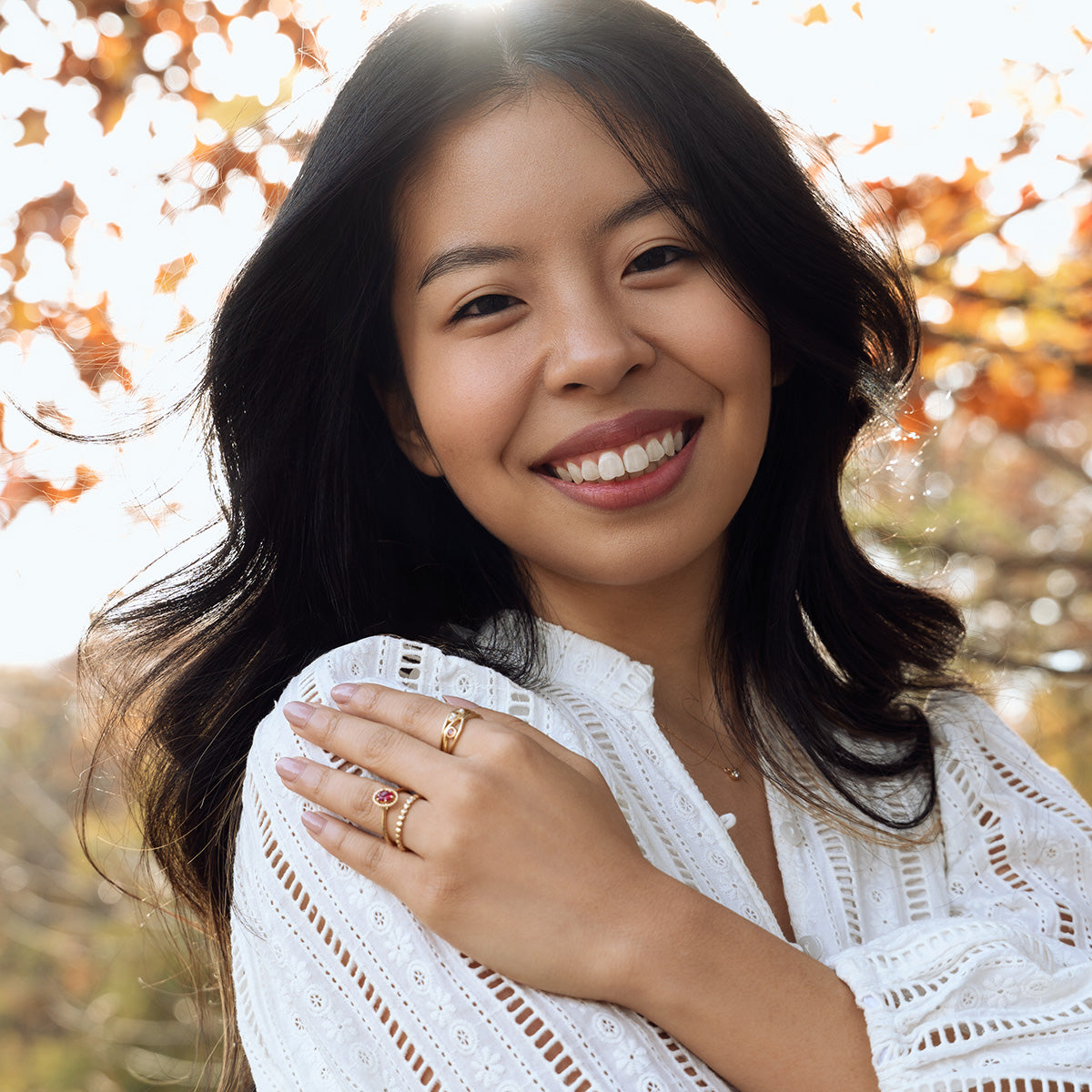 A young woman with long dark hair smiles outdoors, wearing a white blouse and the 10k Hidden Message Morse Code Diamond Band. Sunlight filters through autumn leaves, adding a soft golden glow to the scene.