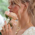 A woman with wavy blonde hair holds pale pink roses near her face, wearing Opal Majesty Studs and a white lace-trimmed top. The softly blurred greenery in the background creates a dreamy atmosphere.