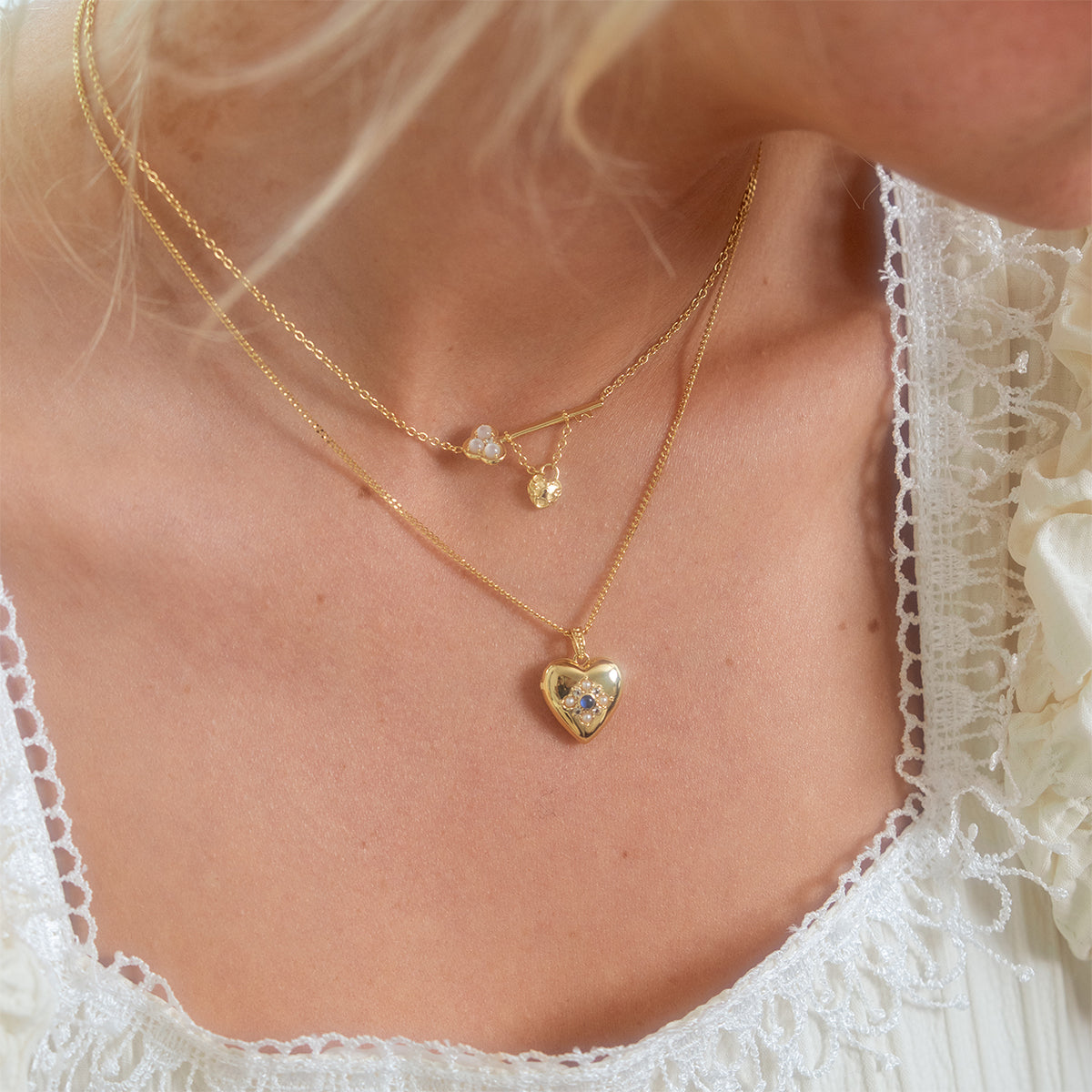 A woman in a delicate white lace dress showcases two layered gold necklaces: one is a vintage romance piece, and the other is the Lock and Key Necklace featuring a gold heart-shaped locket with a sparkling gemstone at its center.