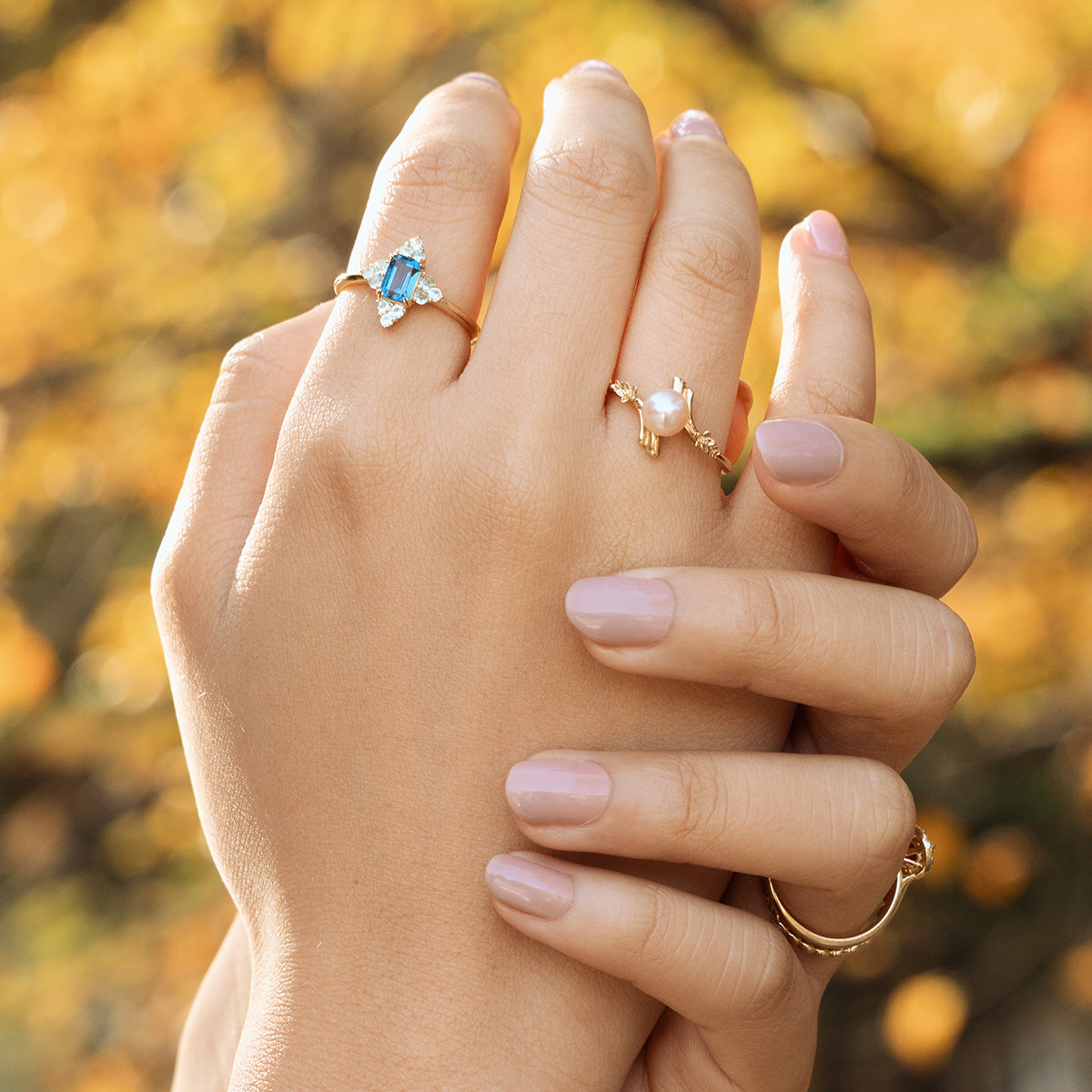 Two hands with manicured nails wearing gold rings, one featuring the 10k Forget Me Not Pink Pearl Bypass Ring, are gently clasped together against a blurred, autumn-colored background.