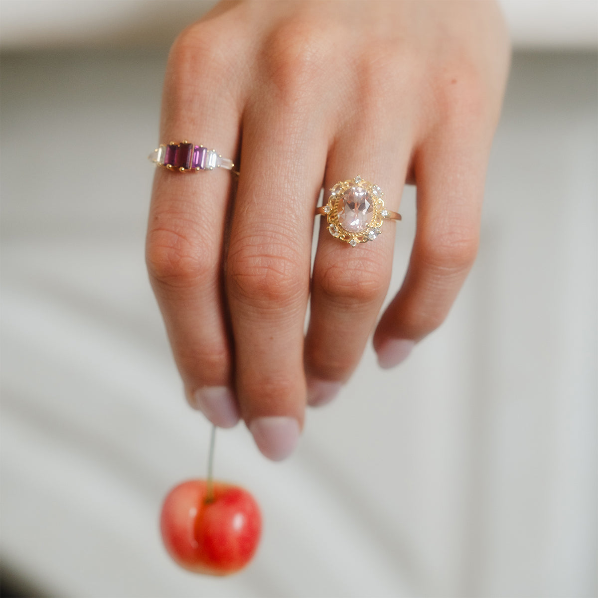 A manicured hand holding a cherry by its stem wears two rings: a 14k gold vermeil band with purple stones and the Royal Blush Cocktail Ring, which features a large oval gemstone in an ornate gold setting.