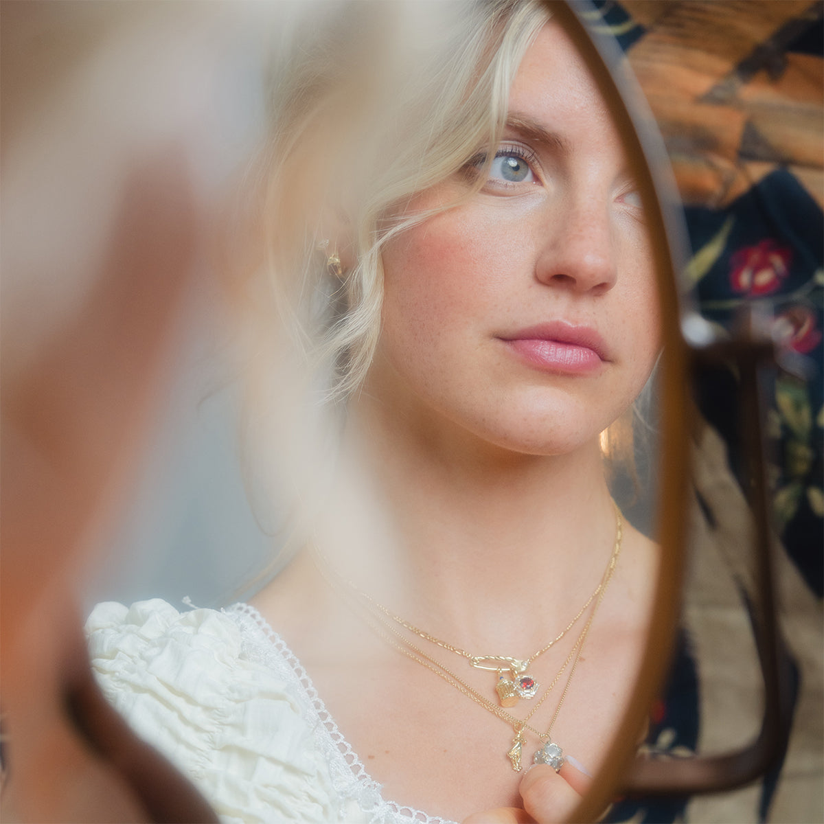 A blonde-haired woman with blue eyes gazes into the mirror, wearing layered gold necklaces, a white ruffled top, and Gilded Garnet Huggie Hoops. Her calm expression is reflected as she admires her elegant gold vermeil earrings.