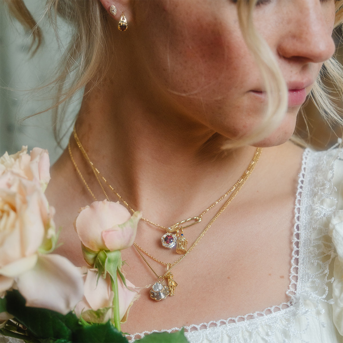 A woman in a white lace dress holds pale pink roses, wearing vintage-inspired jewelry—a gold layered Forget Me Not Blue Topaz Memento Necklace and matching earrings. Her hair is loosely styled, with only part of her face visible.