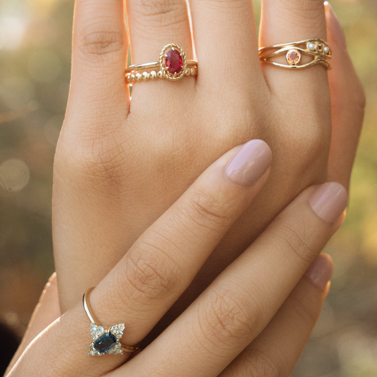 Two hands with light pink manicured nails wear the 10k Hidden Message Morse Code Diamond Band, gleaming with delicate diamonds, alongside other gold rings, set against a softly blurred background.