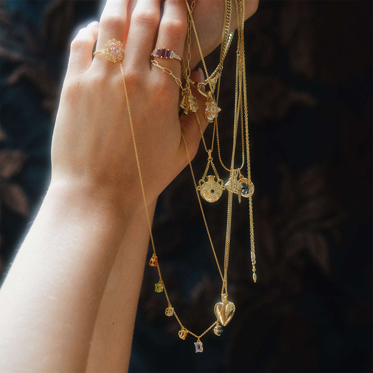 A hand holds several Classical Confetti Gemstone Necklaces in 14k gold vermeil with various pendants, including hearts and gemstones, and wears gold rings with light-colored stones against a dark, blurred background.