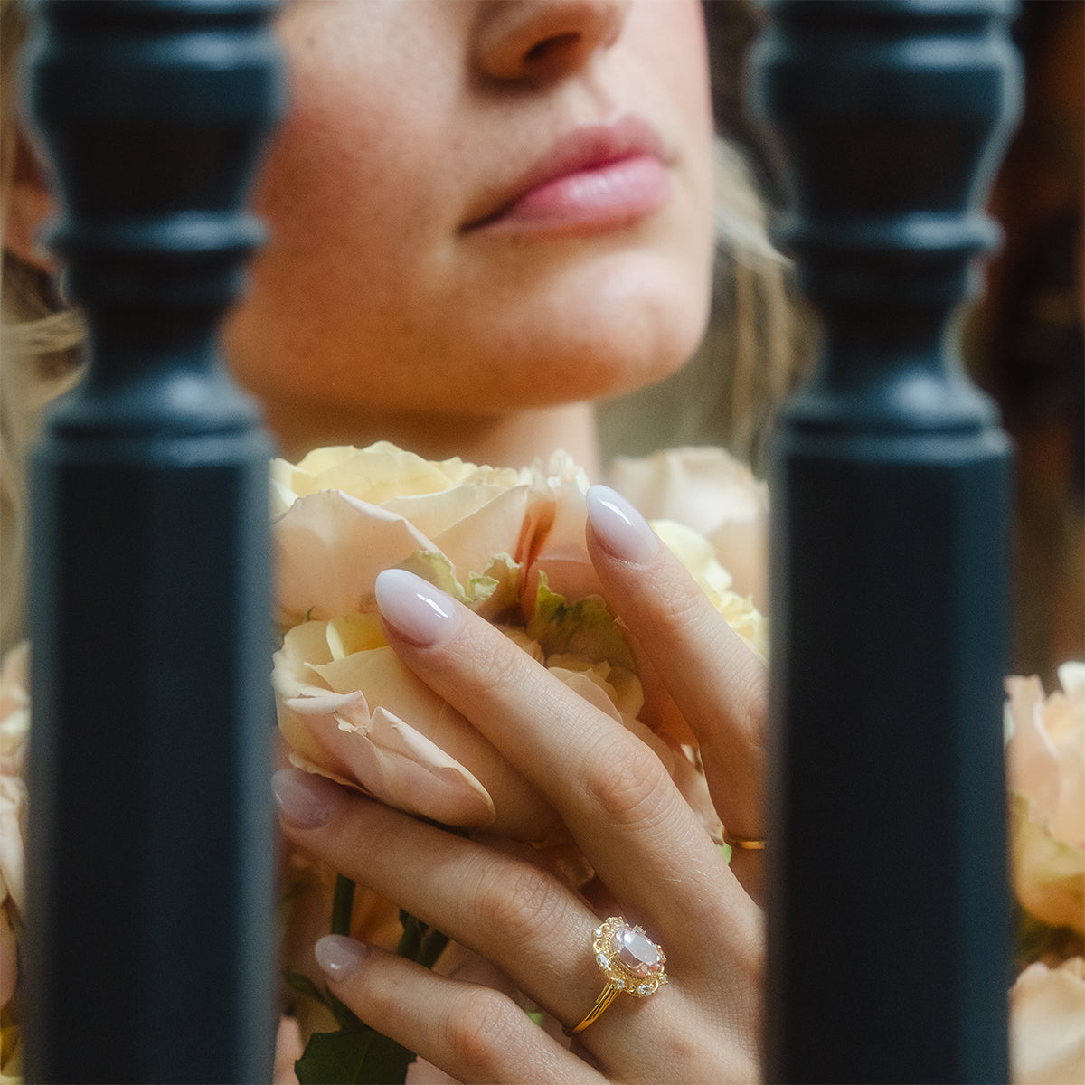A woman with manicured nails holds pale roses near her face, wearing the Royal Blush Cocktail Ring—a 14k gold vermeil piece with a large, light gemstone. Two black bars appear in the foreground.