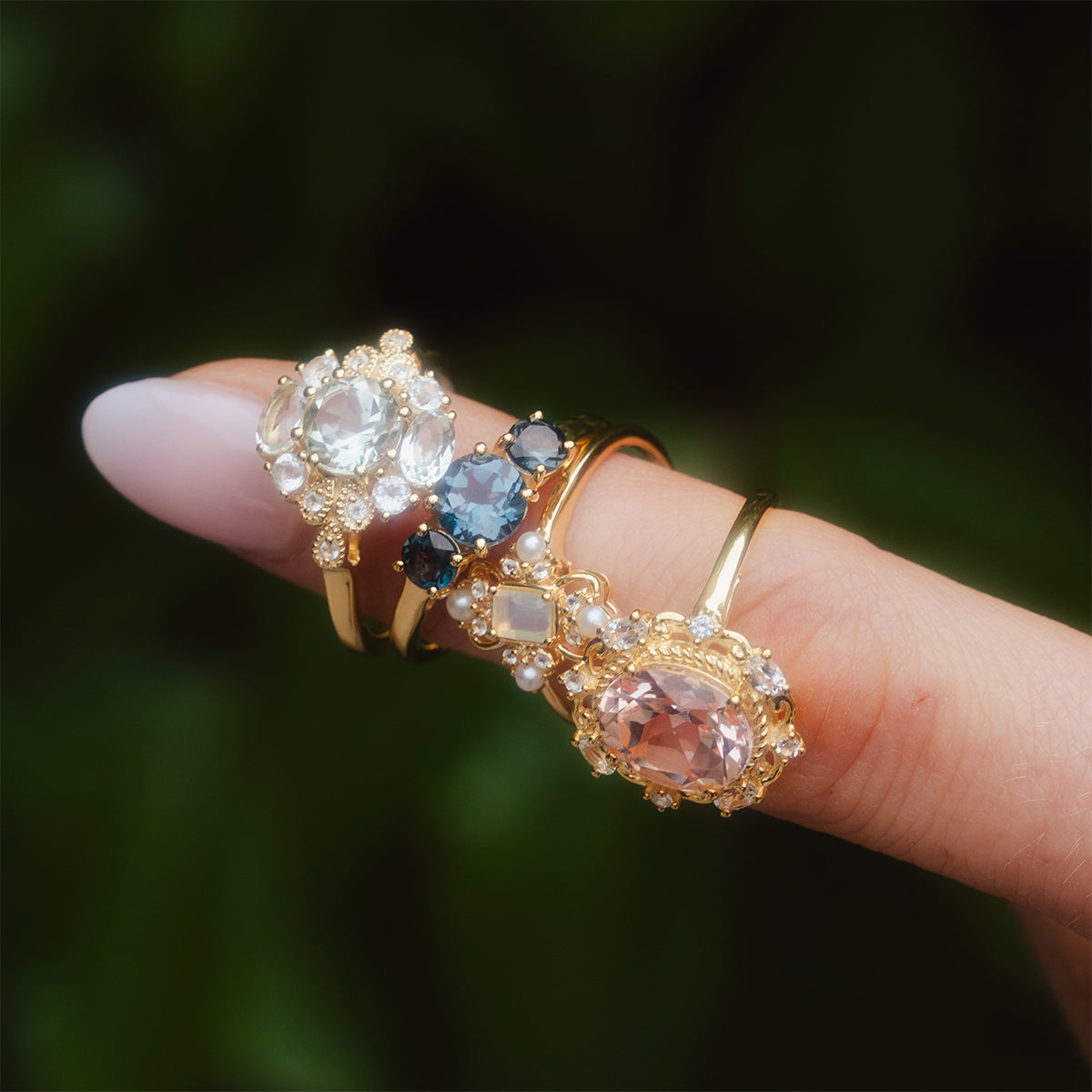 A hand with light pink nail polish displays the Royal Gardens Green Amethyst Cocktail Ring in 14k gold vermeil, featuring a large round green amethyst surrounded by smaller stones, against a blurred green background.