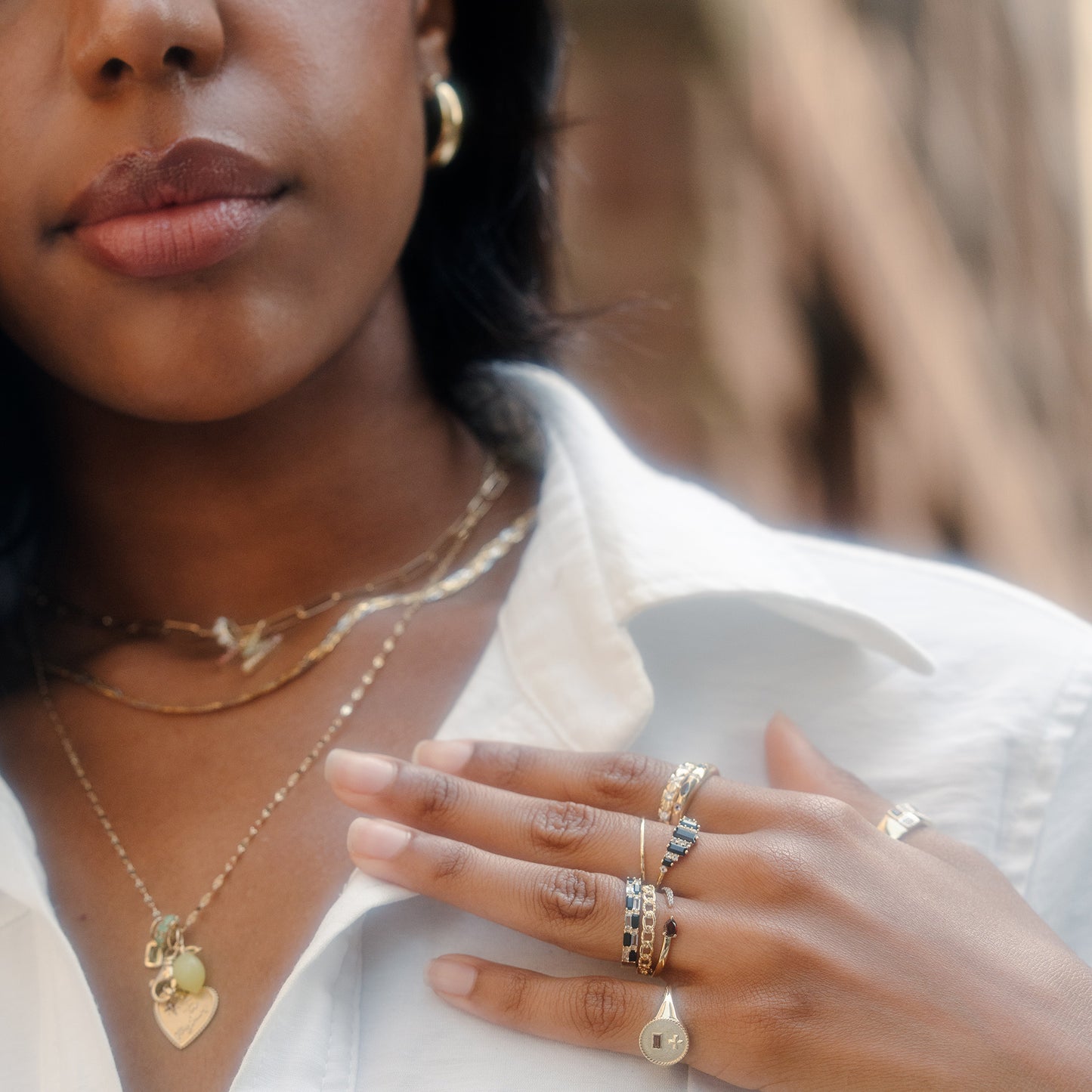 A woman in a white shirt displays layered 9k yellow gold necklaces, one with a heart pendant, and wears the Solid Gold Garnet and Sapphire Open Front Band among other rings. Only her lower face, neck, and hand are visible.