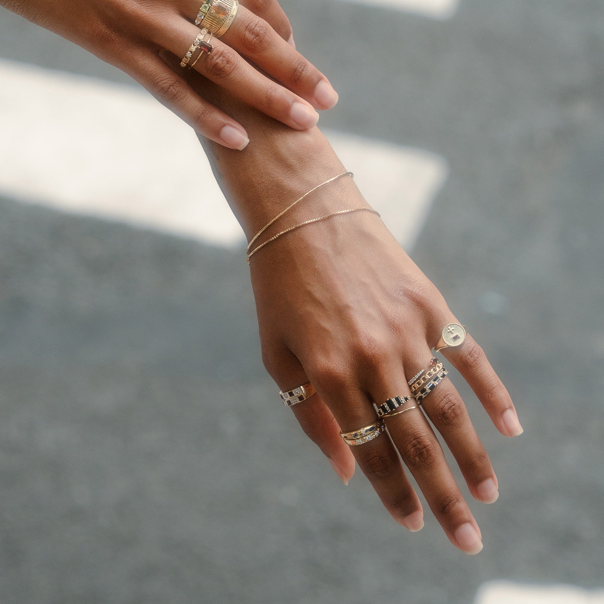 A close-up of two brown-skinned hands adorned with several gold rings, including the Solid Gold Pizza Acrostic Ring, and a delicate bracelet, gracefully positioned against a blurred gray backdrop.