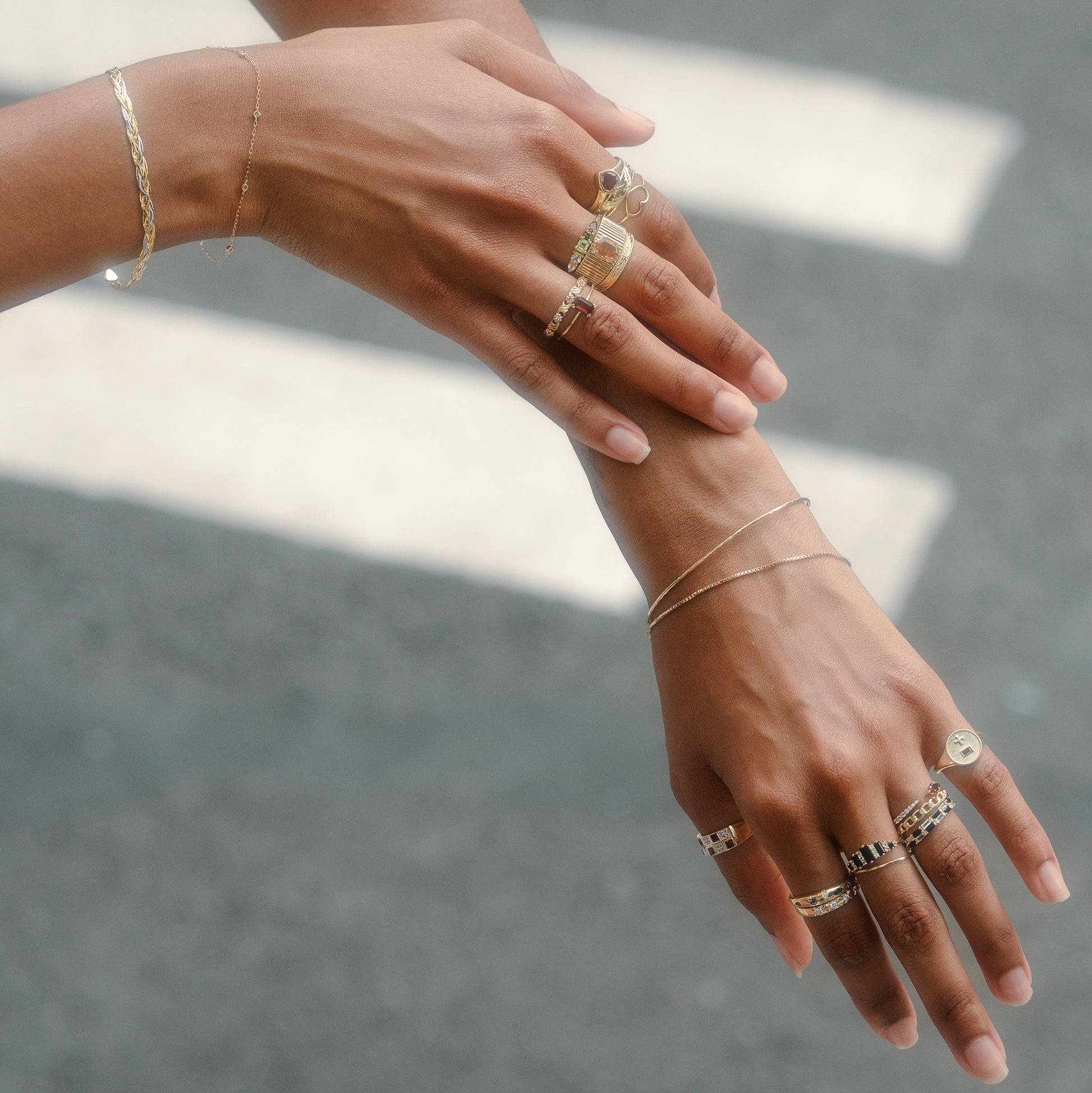 Two hands with light brown skin wear multiple gold rings and the Solid Gold Structured Bracelet, along with delicate bracelets and a chain bracelet, all displayed gracefully against a blurred gray background with white lines.