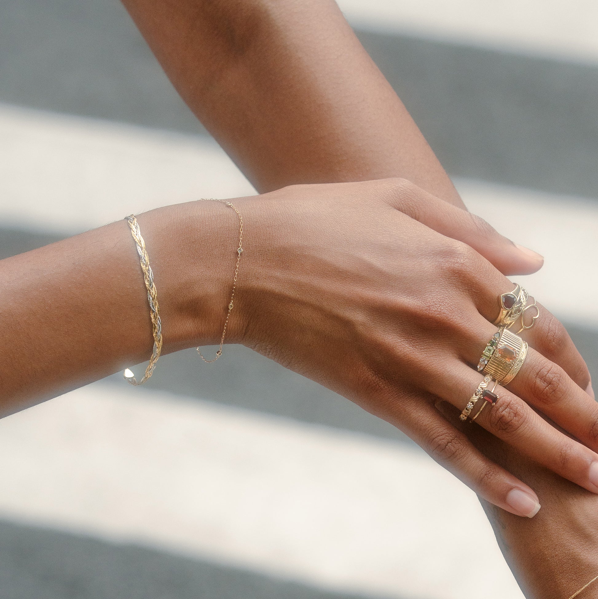 Two brown-skinned hands adorned with gold rings and a Solid Gold Mixed Metal Braided Bracelet gently touch, set against a blurred backdrop with white stripes reminiscent of a crosswalk.