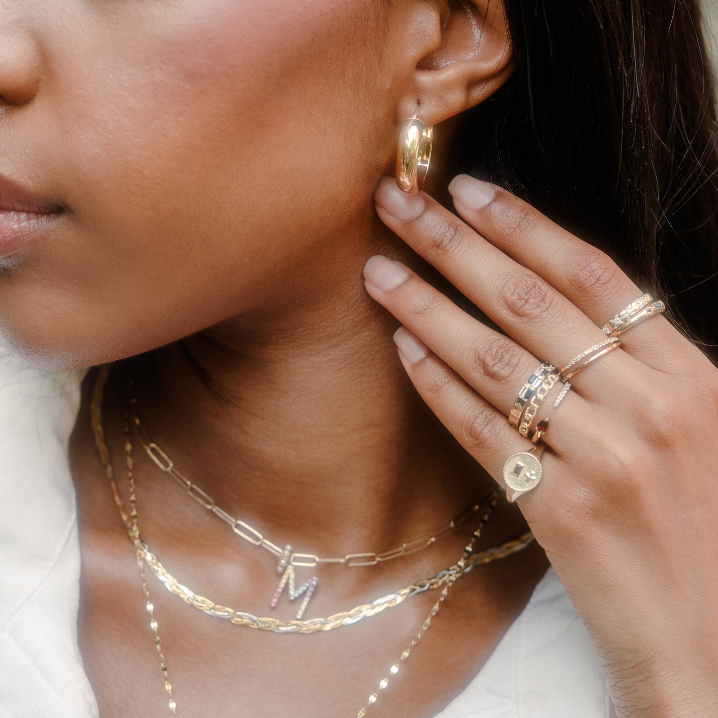 A close-up of a woman wearing 9k yellow gold jewelry, including hoop earrings, layered necklaces with an "M" pendant, and the Solid Gold Garnet and Sapphire Open Front Band on her finger, against a softly focused neutral background.