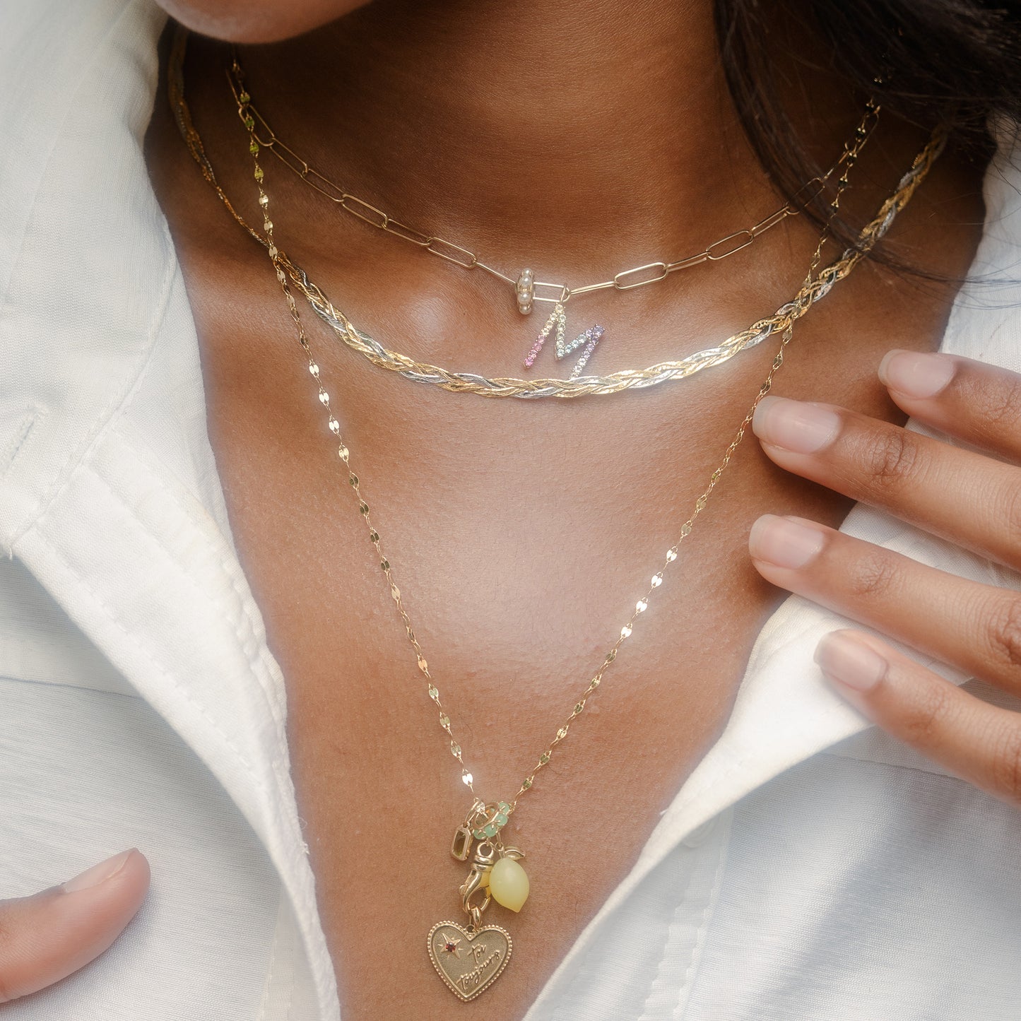 A close-up of a person wearing a white shirt and the Solid Gold Pastel Gemstone Letter Charm necklace, featuring layered gold chains with a heart pendant and pastel gemstone jewelry. Their hands gently touch their collar.