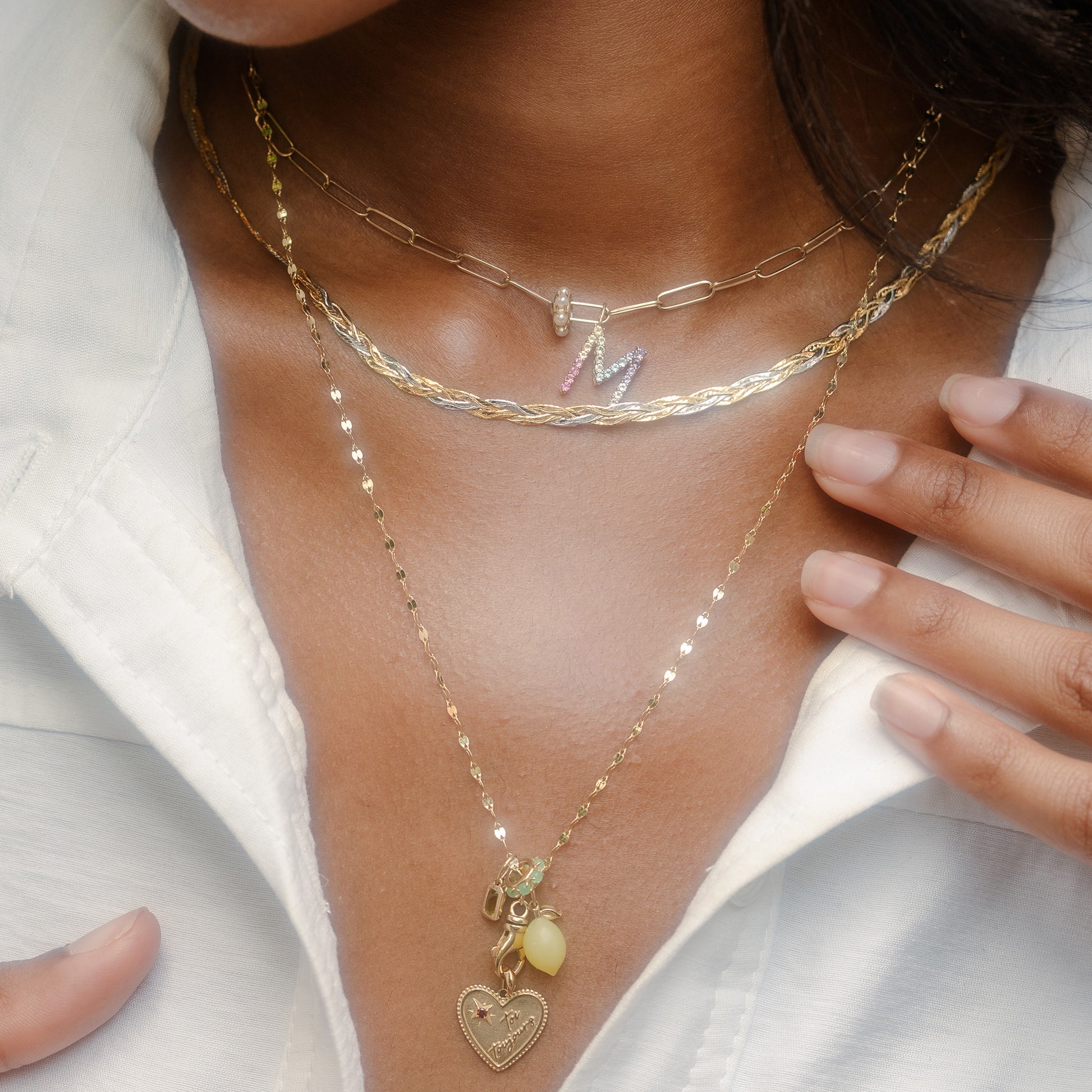 A close-up of a person wearing a white shirt and the Solid Gold Pastel Gemstone Letter Charm necklace, featuring layered gold chains with a heart pendant and pastel gemstone jewelry. Their hands gently touch their collar.