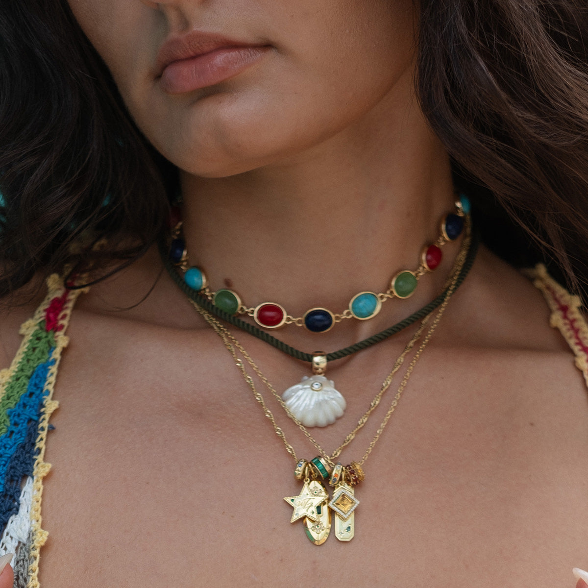 A woman wears a 14k yellow gold plated charm necklace: a colorful beaded choker, a gold chain with shell pendant, and a longer chain featuring stars, gems, and the Hidden Curse Word Spacer Charm. She pairs it with a multicolored crocheted top.