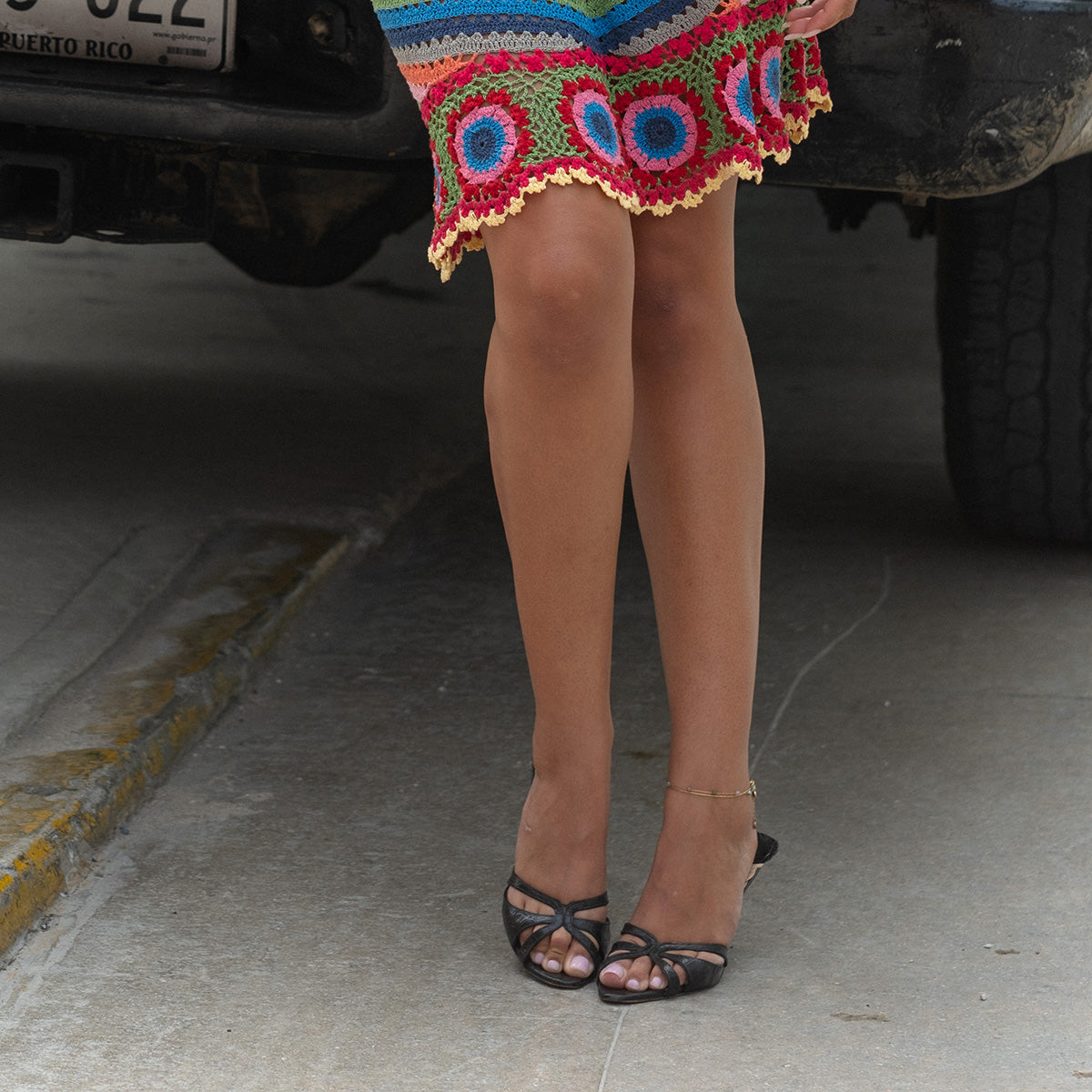 A person wearing a colorful crocheted skirt with circular patterns and black heeled sandals, styled with the Alexis Anklet, stands on a concrete surface near a parked vehicle.