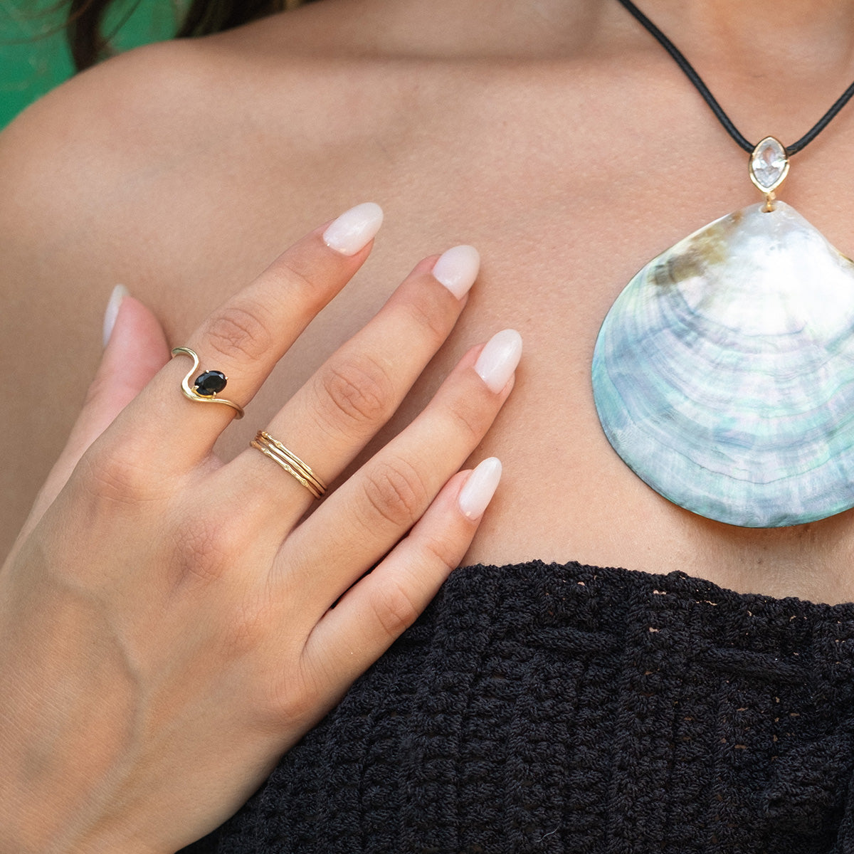 A close-up of a woman's hand with natural nails, wearing gold rings and the Solid Gold Dainty Birthstone Stacking Ring, rests near a large shell pendant necklace. She is dressed in a black textured top.