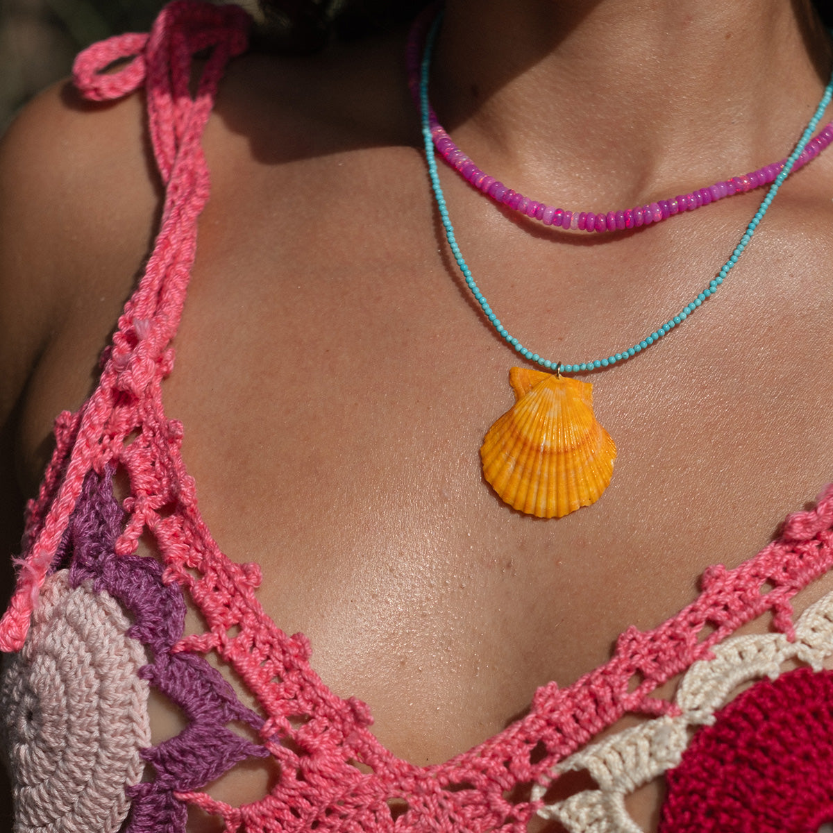 A person wears a colorful pink and cream crochet top and two necklaces—one with purple beads, the other is the Byron Necklace featuring a seashell accent. The photo is taken outdoors with sunlight highlighting their skin.