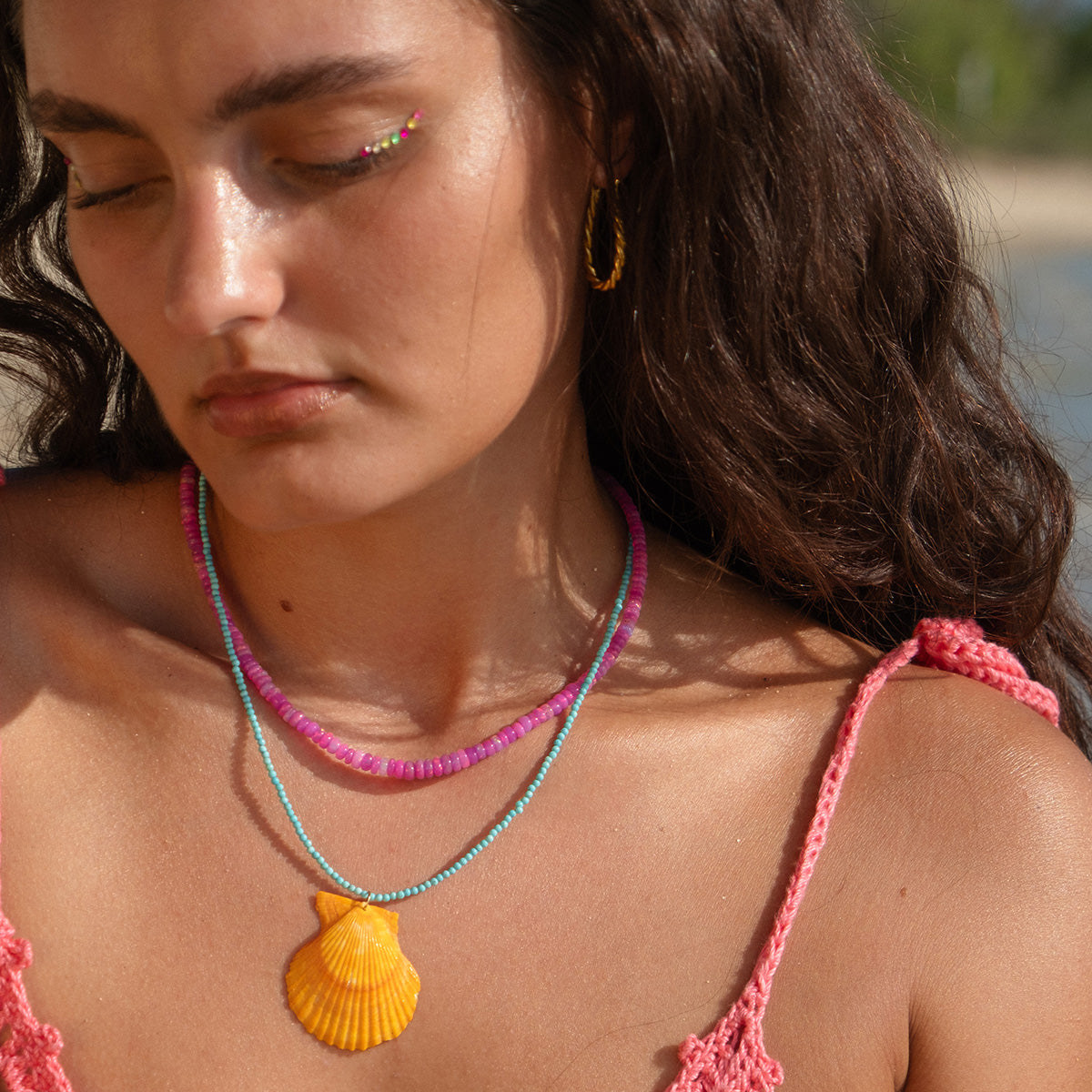 A woman with wavy brown hair wears a pink crocheted top, colorful necklaces, rainbow glitter eyeliner, and the Twisted Oblong Hoops. She is outdoors near the beach.