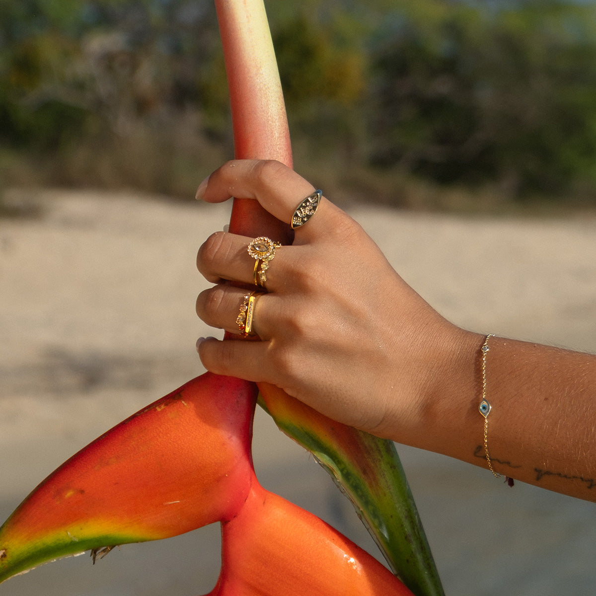 A hand wearing the Colorful Language Fuck Signet Ring and a 14k gold plated bracelet holds a vibrant orange and green tropical flower outdoors, with sand and greenery blurred in the background.