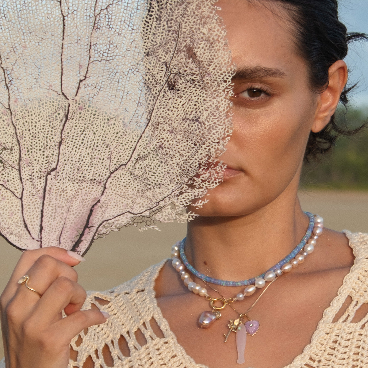 A woman stands outdoors holding a sea fan in front of half her face, wearing layered Luiny x Local Eclectic Charms necklaces, a handmade pendant, and a gold ring against a blurred sandy background.