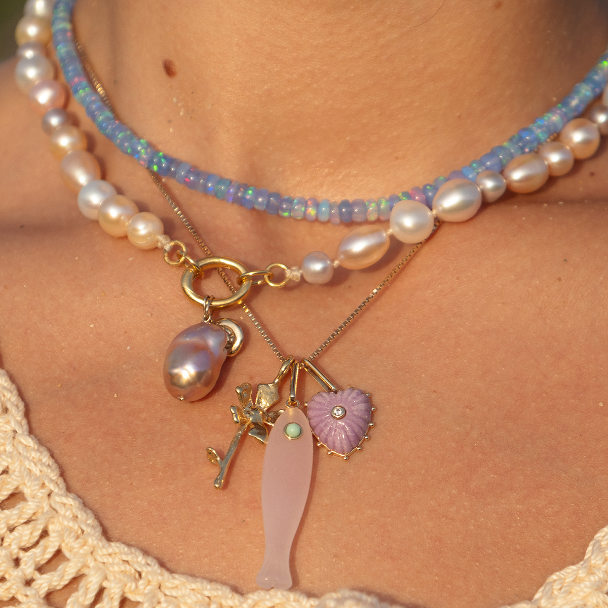 Close-up of a neck adorned with layered Luiny x Local Eclectic Charms necklaces: pearls and blue beads, a large pearl strand, and a gold chain with solid brass pink fish, pink heart, and green stone branch charms.