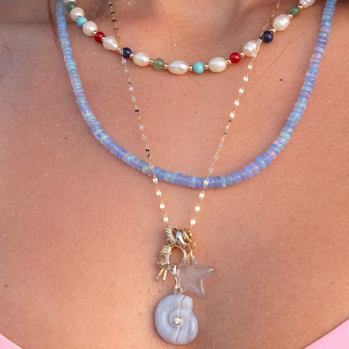 Close-up of a person's neck wearing three layered necklaces: a colorful beaded necklace with pearls, a blue beaded necklace, and a Family Gold chain featuring star, Solid Gold Body Language Charm (astronaut), and round blue gold charm pendants.