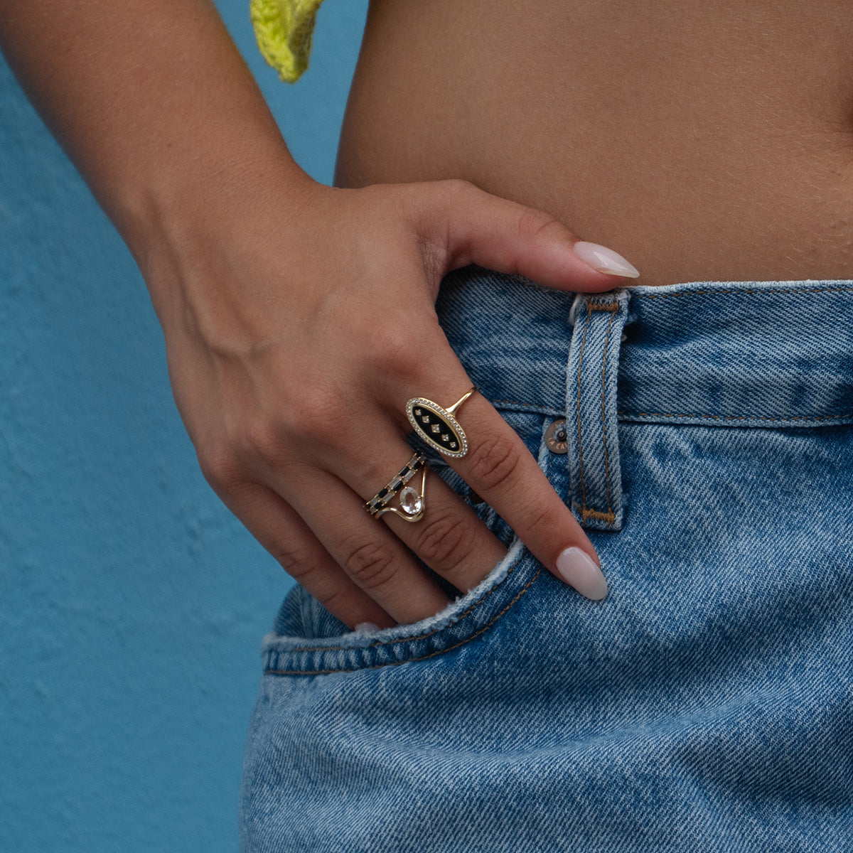A person wearing denim jeans rests their hand in a pocket, displaying stylish rings—including the Solid Gold Black and White Checkerboard Band. A light blue wall and a hint of yellow top are seen in the background.