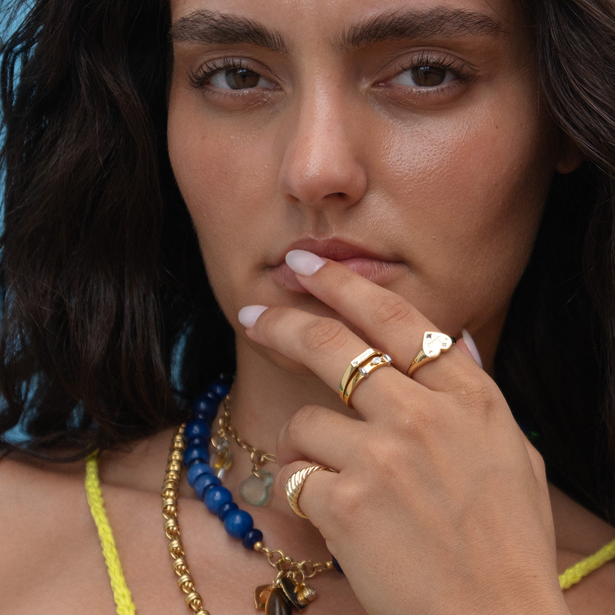 Close-up of a woman with wavy dark hair wearing the Solid Gold Twisted Signet Ring, layered blue-beaded necklaces, and a yellow strap top. She touches her lips thoughtfully, radiating calm confidence.