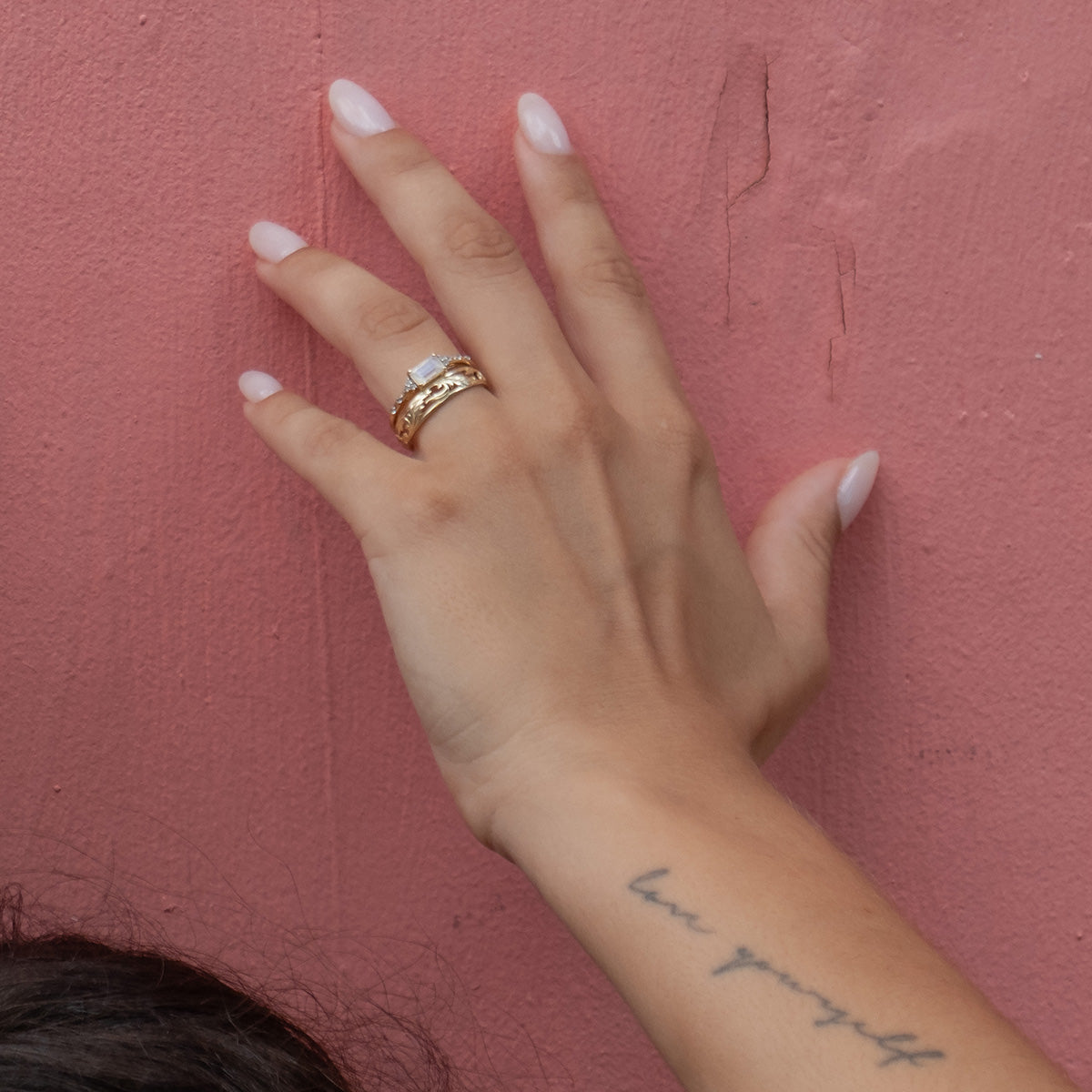 A hand with a light pink manicure, wearing the Solid Gold Glimmering Birthstone Baguette Ring and another gold band, rests against a textured pink wall. A script tattoo reading "love yourself" graces the wrist.