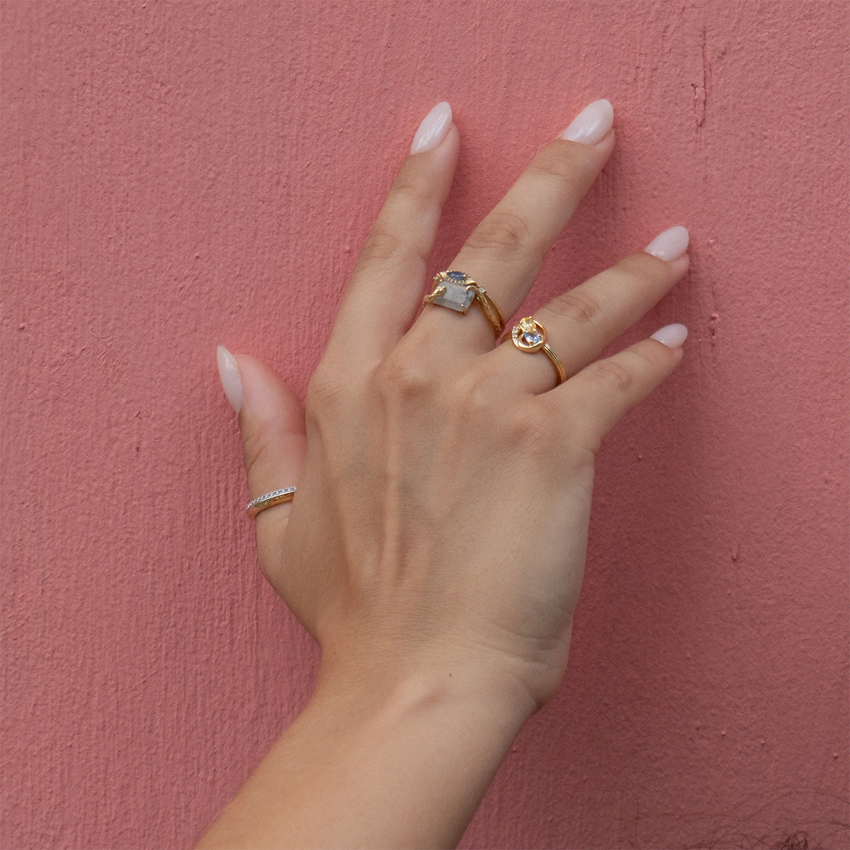 A hand with light pink nail polish rests on a textured pink wall, wearing three rings: a slim band on the pinky, the Solid Gold Labradorite Serpent Ring for Protection on the middle finger, and a gold ring with a round stone on the ring finger.