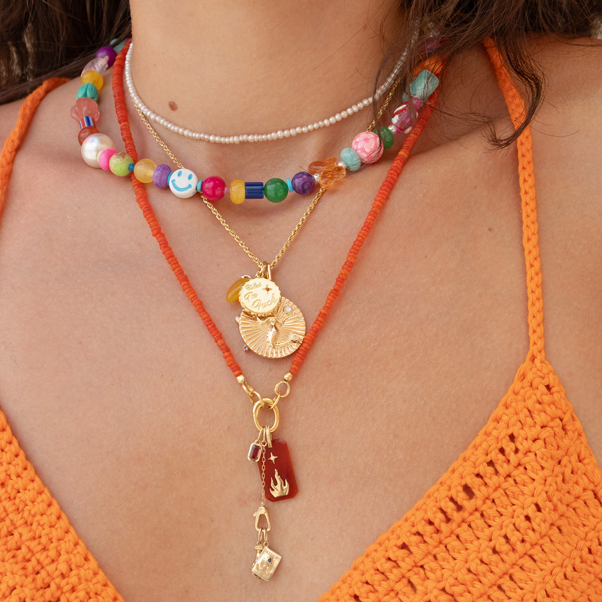 A person wearing layered necklaces, including a colorful choker, a thin pearl necklace, gemstone statement necklaces with gold coin pendants, and the Beaded Gemstone Charm Holder Necklace in orange, paired with an orange crochet top.