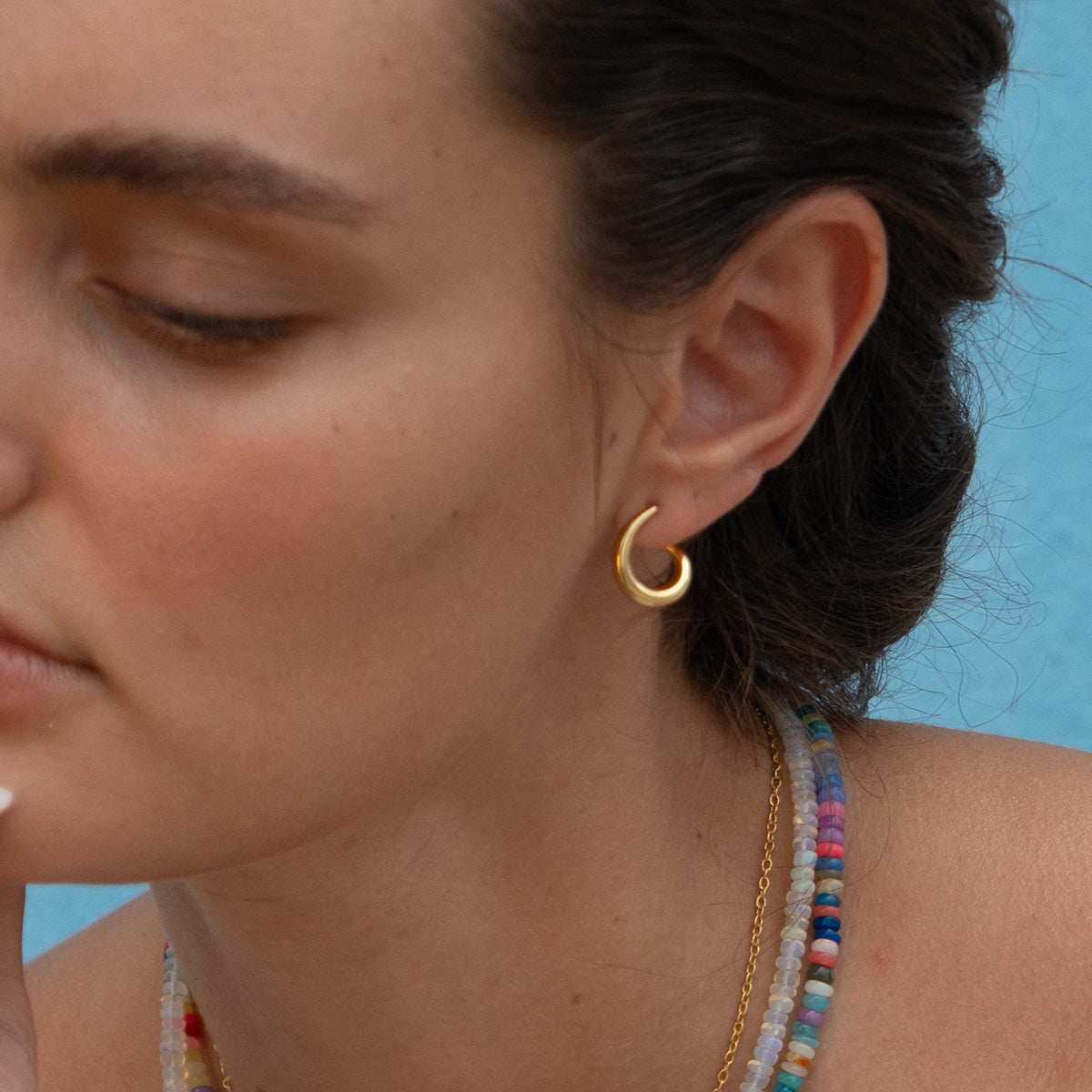 A light-skinned woman with brown hair wears the 14k gold-plated Modern Loop Hoop Earrings, styled with layered colorful beaded and gold chain necklaces, set against a blue background. She poses calmly and contemplatively.