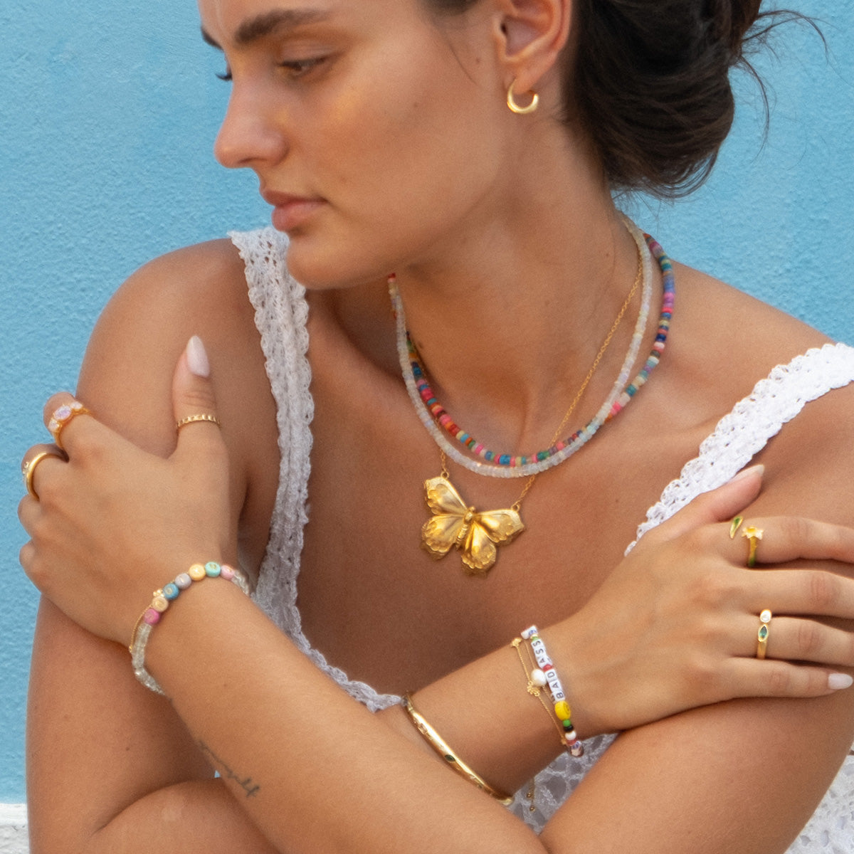 A woman with dark hair in a white lace top poses against a blue wall, wearing colorful beaded necklaces, gold earrings, multiple gold rings and bracelets, and a striking Butterfly Necklace featuring engraved, gold-plated wings.