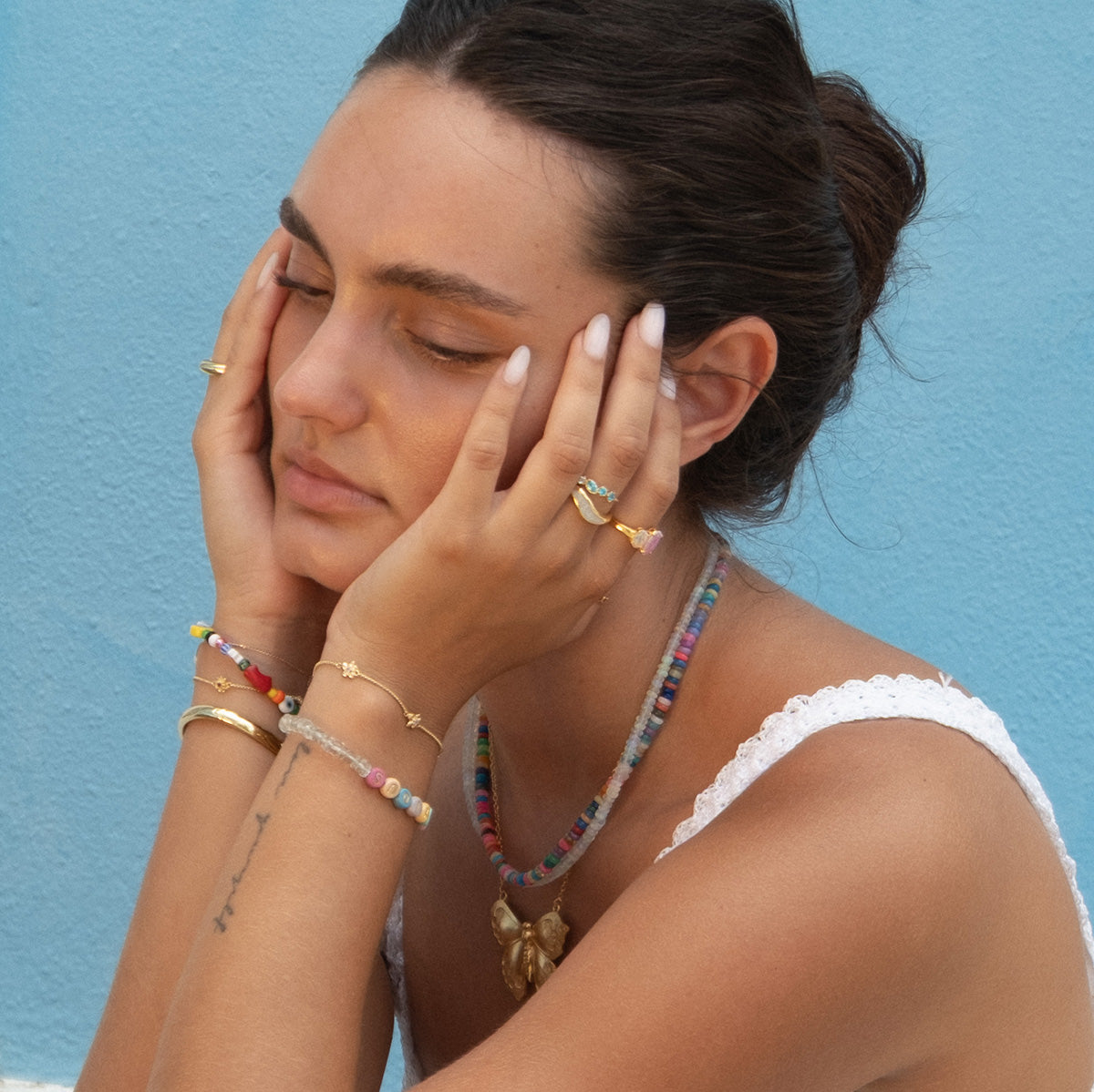A woman with dark hair in a bun, eyes closed, rests her face in her hands, wearing colorful beaded jewelry and rings. She showcases the Butterfly Necklace with engraved wings and gold plating against a light blue wall.