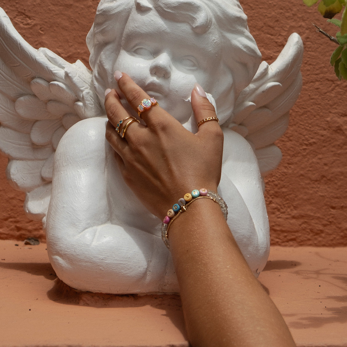 A hand wearing the Beaded Feelings Bracelet and colorful rings with heart charms touches the chin of a white angel statue in front of a peach-colored wall.