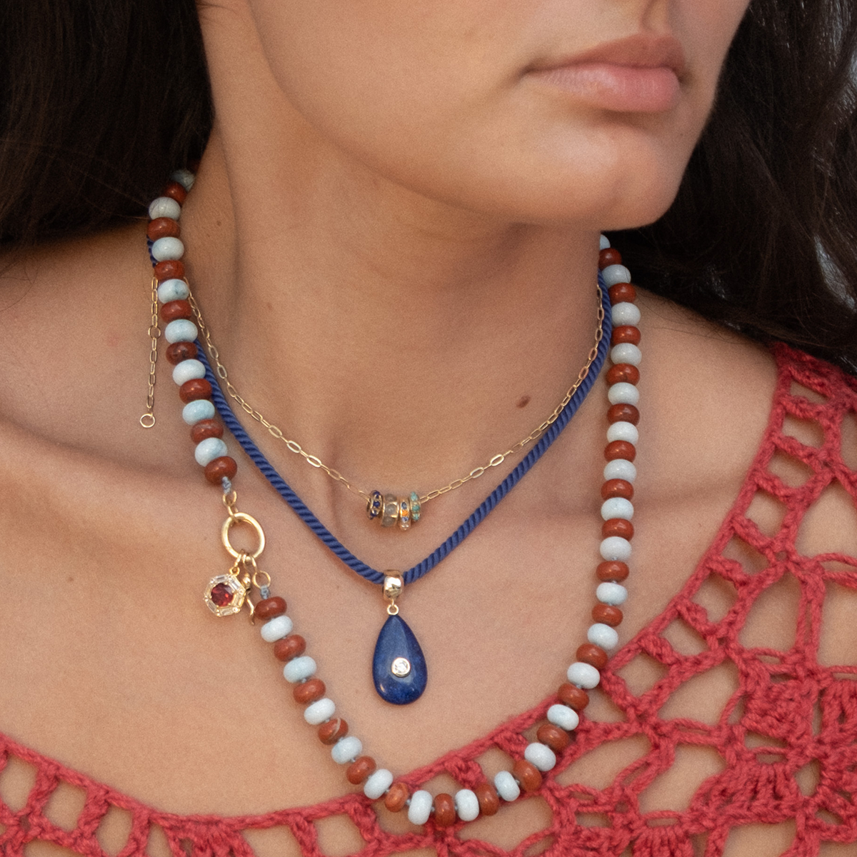 A woman wears the Charms by Thatch: a red crocheted top and three layered necklaces—a beaded necklace with 14k yellow gold plated charms, a gold chain with small beads, and a blue cord with a teardrop-shaped blue pendant. Only her lower face is visible.