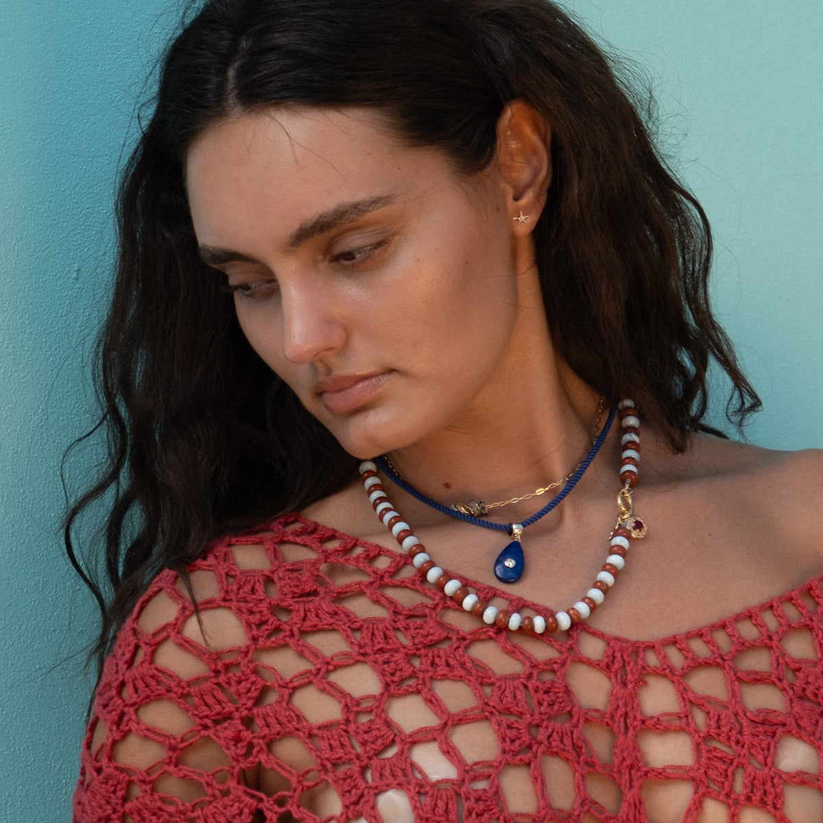 A woman with long dark hair wearing Shimmering Starfish Studs and a pink crocheted top stands against a light blue background, looking down with a serene expression.
