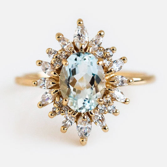 The Lana Ring with Aquamarine & CZ features a large oval aquamarine center, encircled by marquise and round CZ stones in a starburst halo, all set in 14k solid gold against a white background.